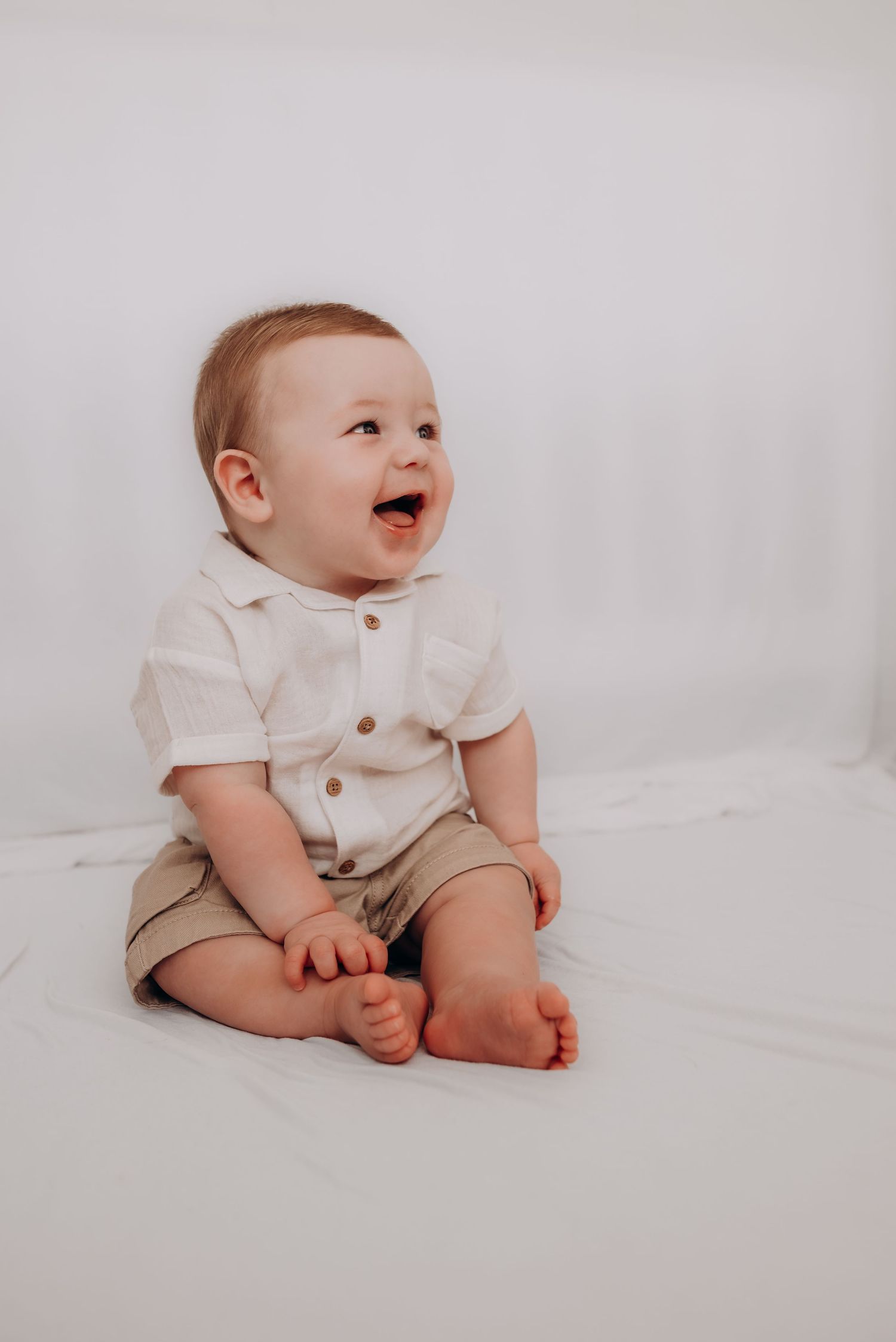 A happy baby in white shirt and khaki shorts sits on white backdrop.