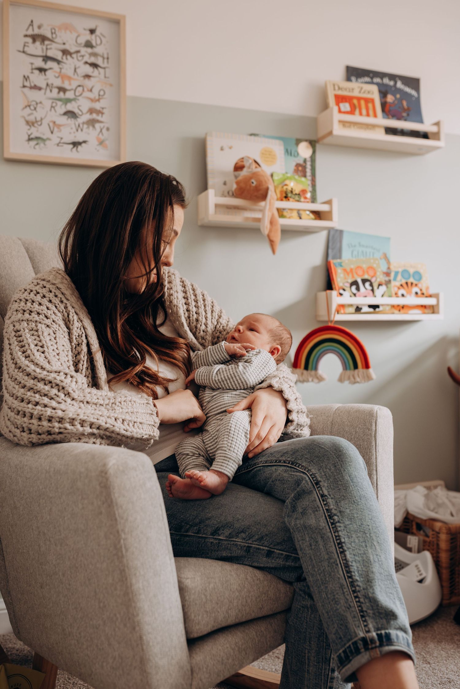 Parent in cozy sweater cuddles with baby on grey sofa surrounded by children's books and decor.