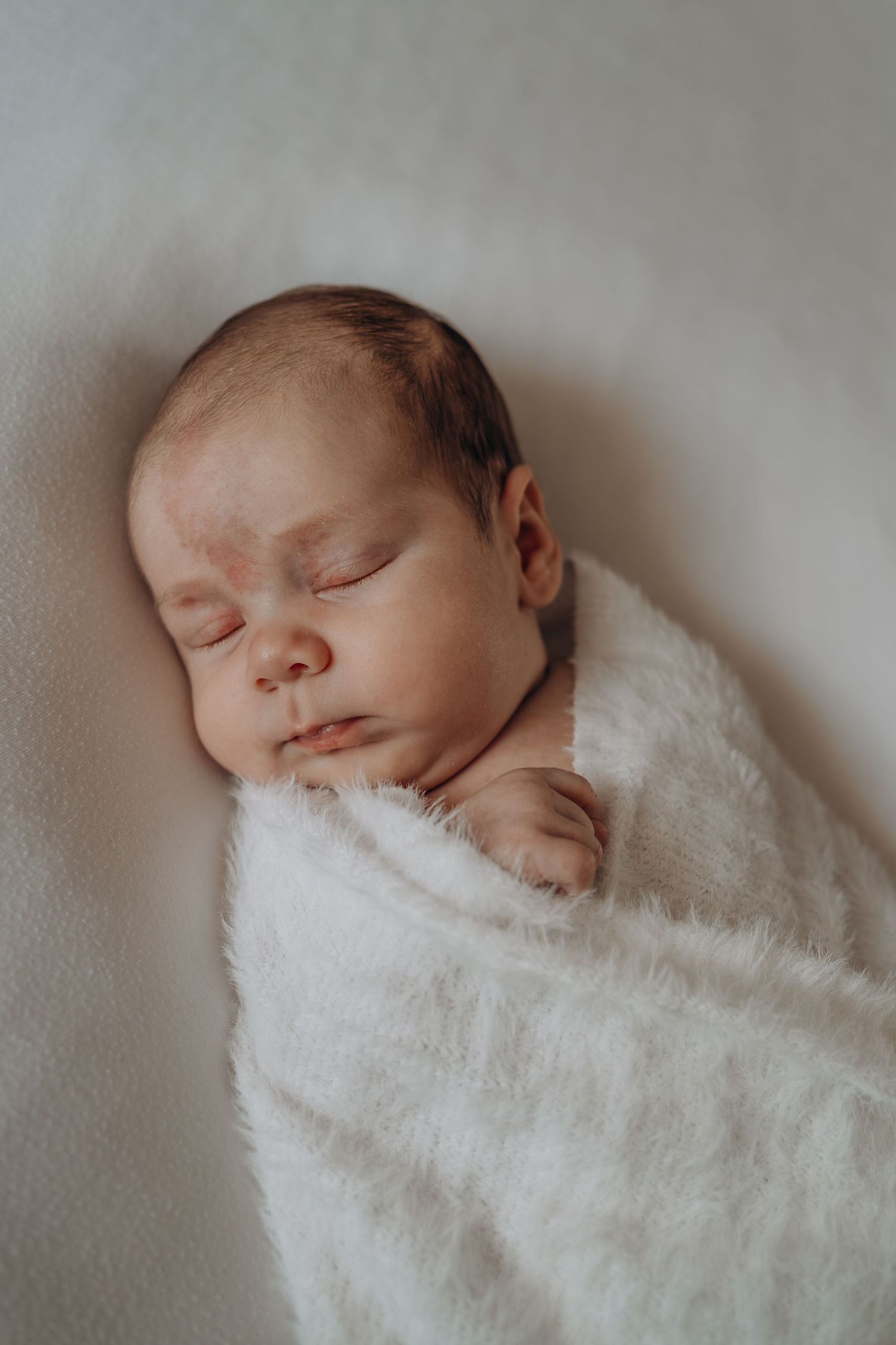 Peaceful newborn baby sleeping swaddled in a soft white fuzzy blanket on a white surface.
