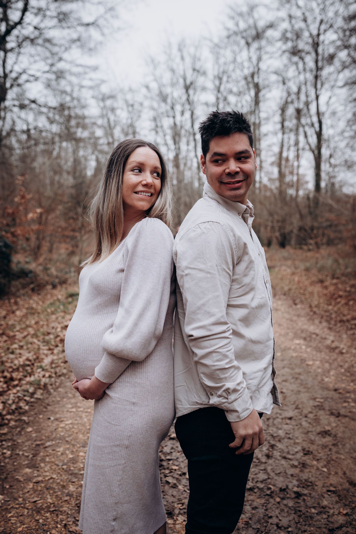 Expectant couple poses back-to-back on an autumn forest trail wearing light colored clothing.