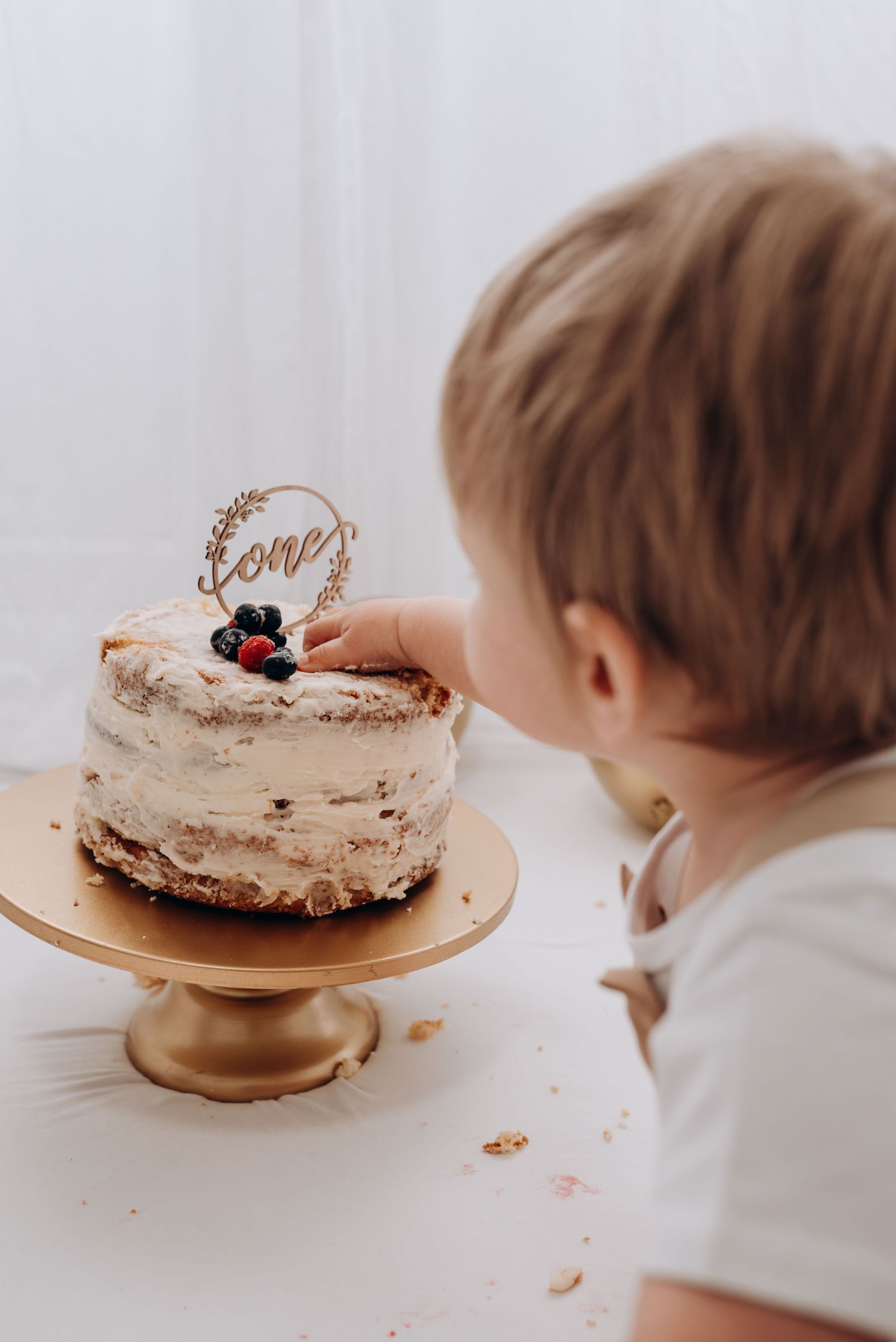 Small child reaching for cake displayed on gold pedestal.