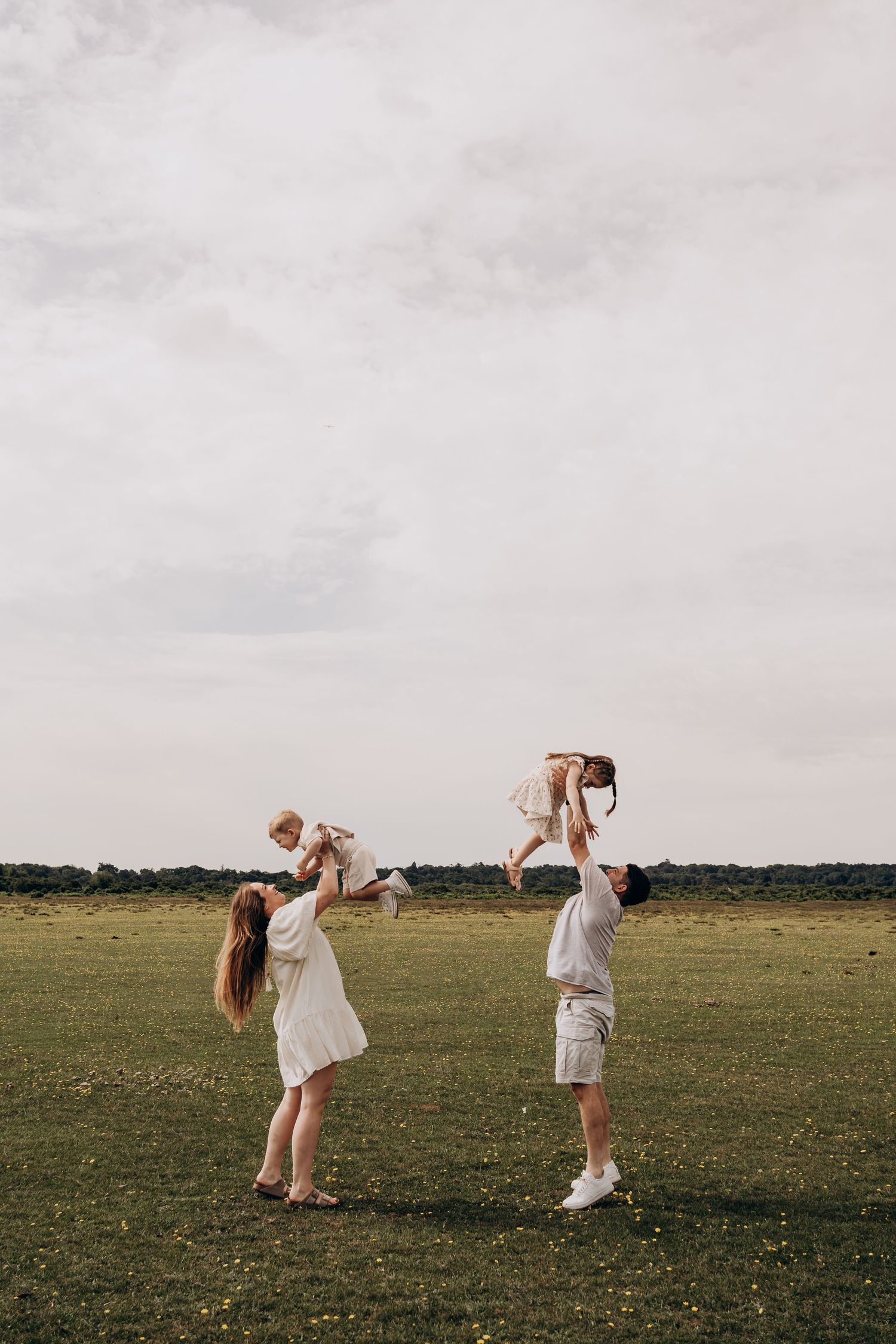 Parents joyfully lift their two young children in a sunny green field under a cloudy sky.