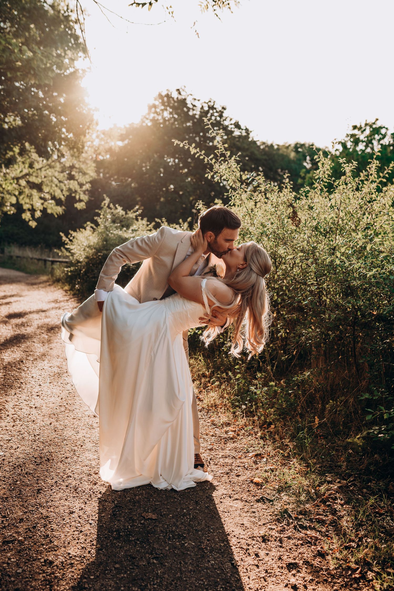 A couple in white outfits dances together on a sunlit path surrounded by lush greenery at golden hour.