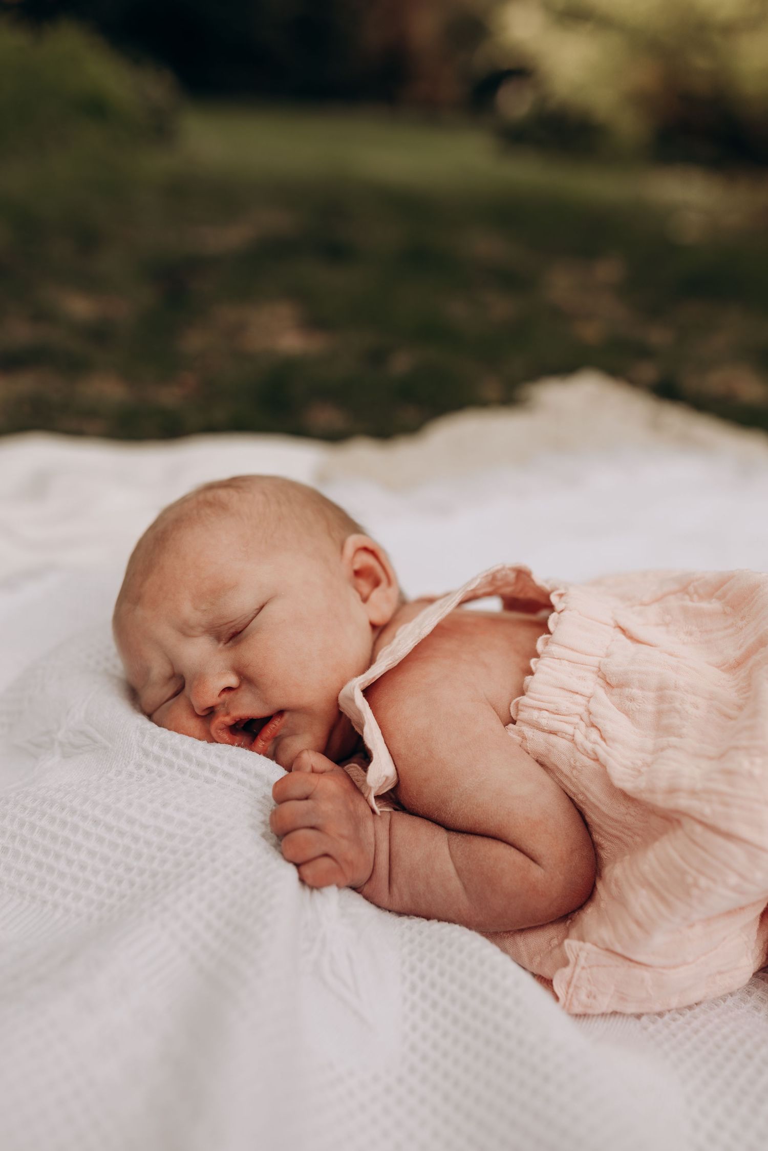 Sleeping newborn baby wearing soft pink clothing rests peacefully on white textured bedding.