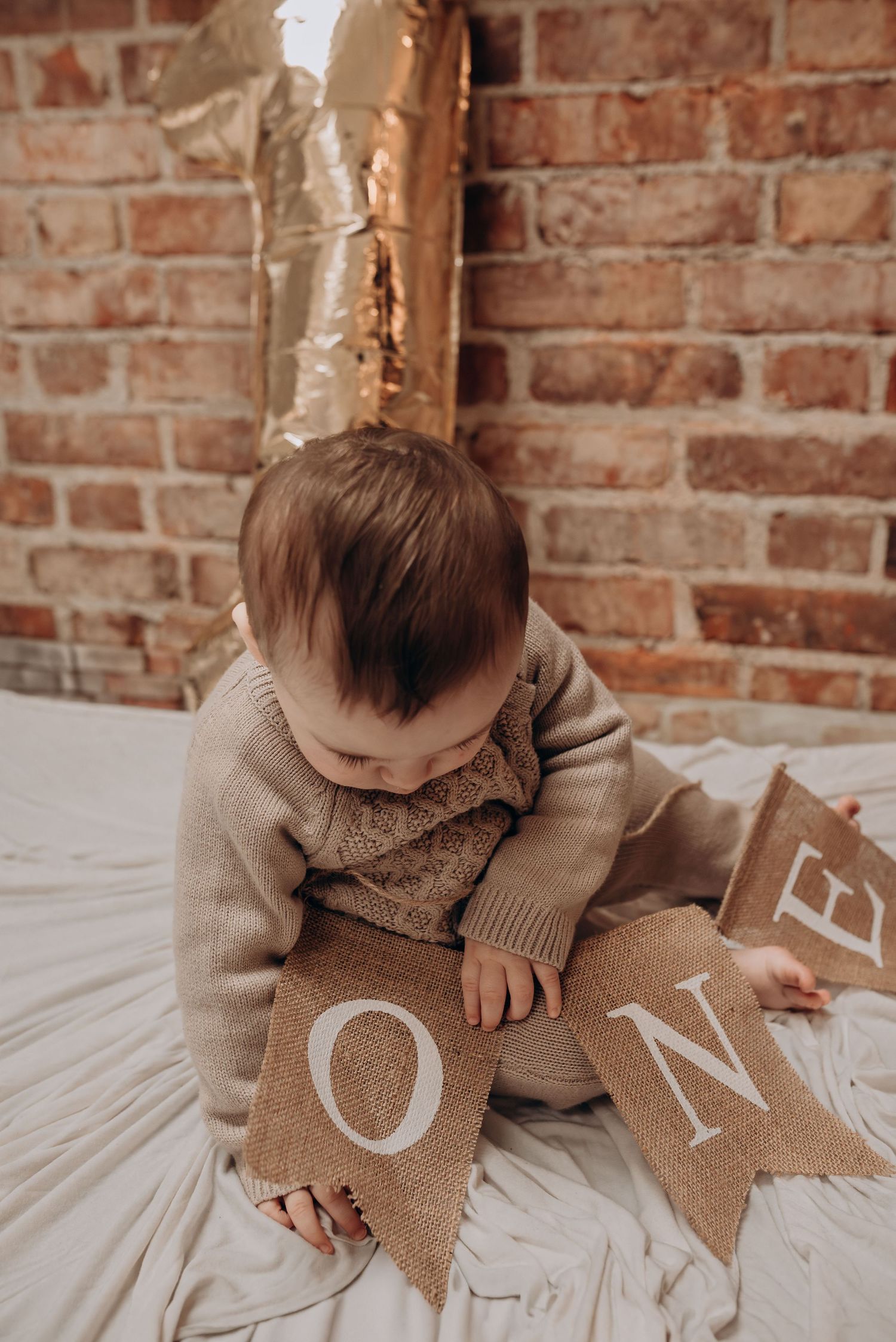 A baby plays with burlap letter decorations against a rustic brick wall background.
