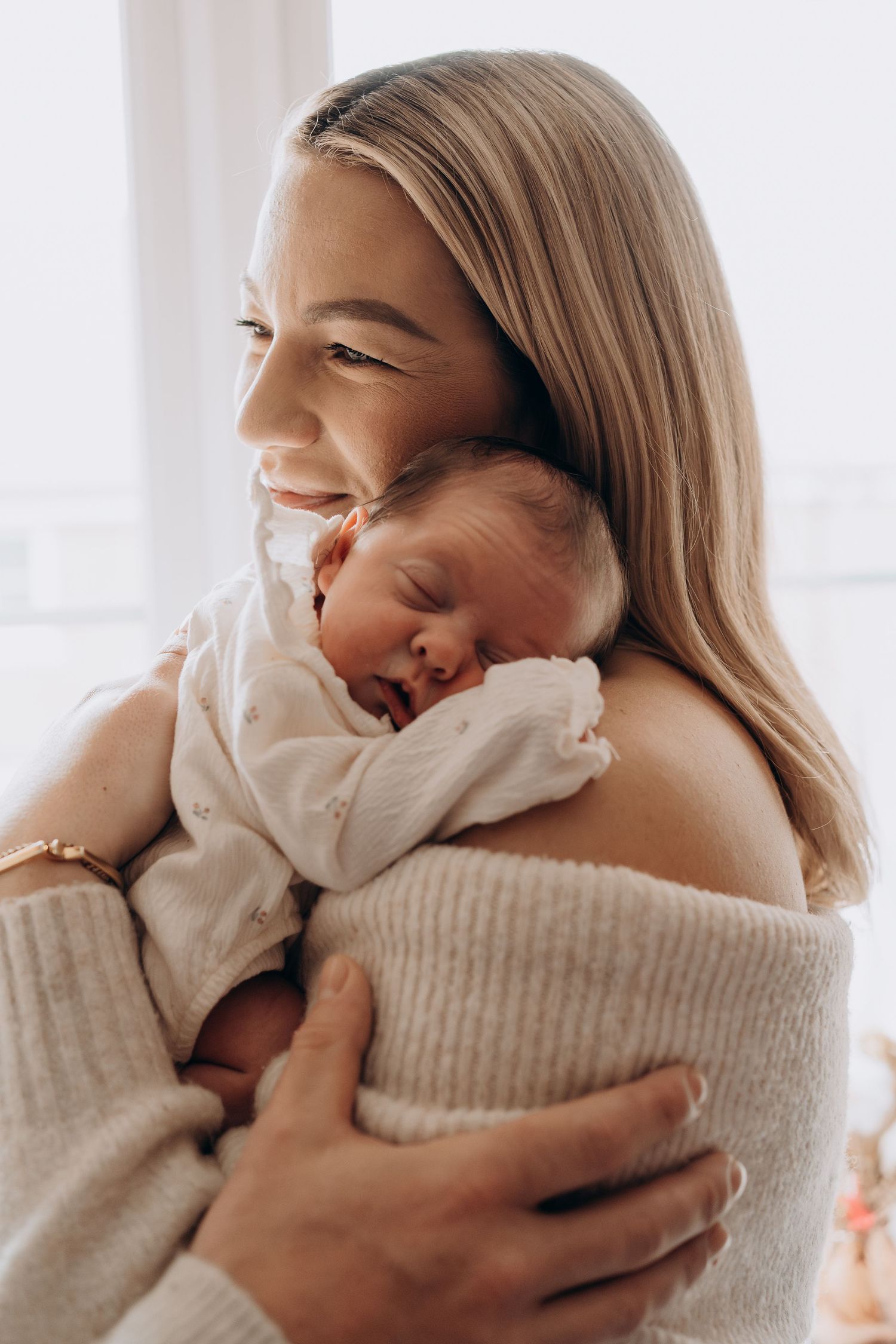 A smiling blonde mother tenderly holds her sleeping newborn baby against her shoulder in a soft, natural light.