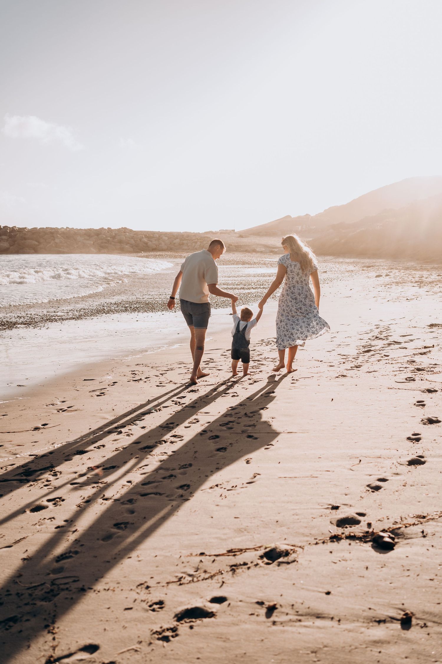 Long shadows cast on beach sand as family strolls along shoreline at sunset.