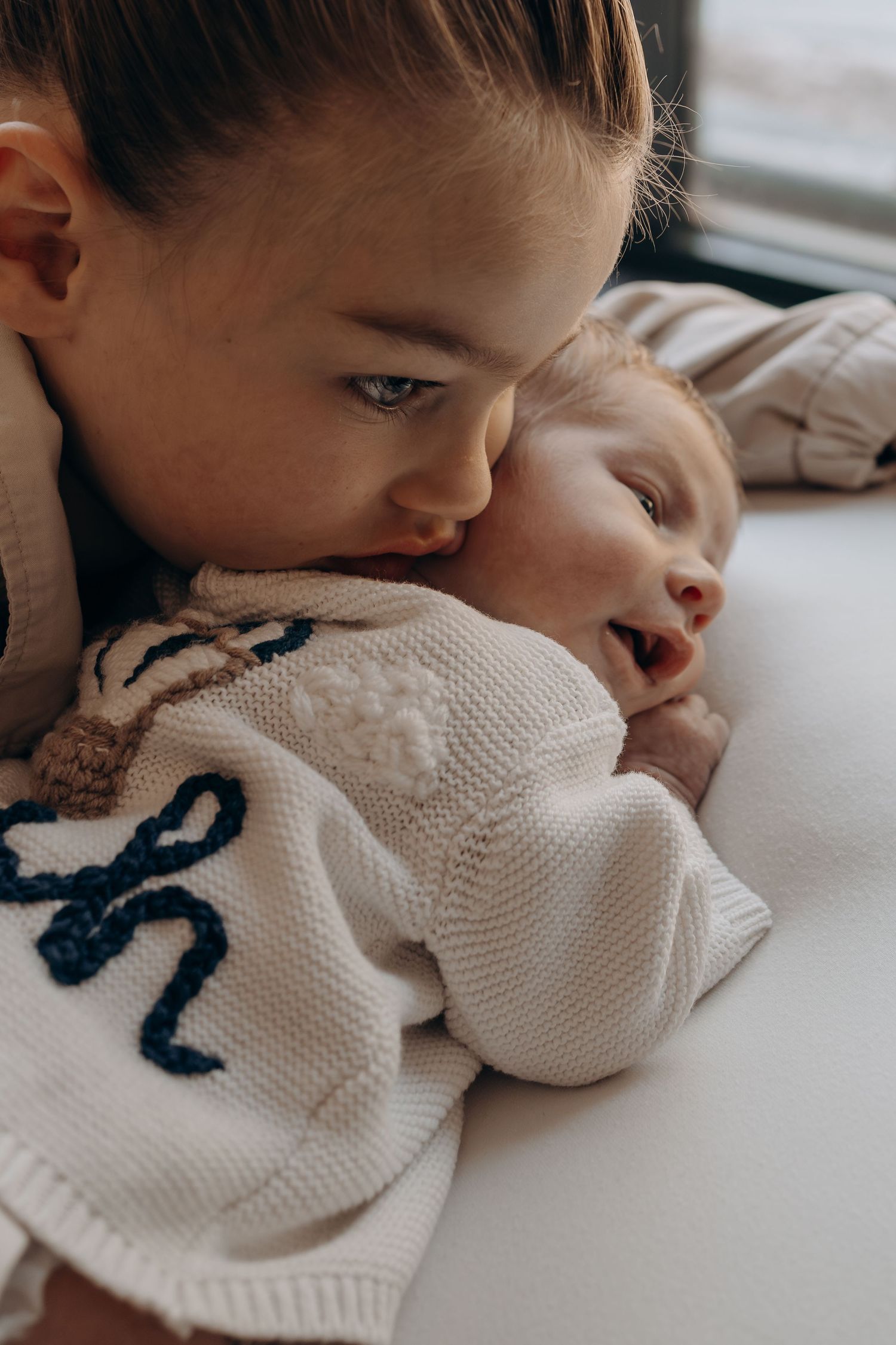 Young girl tenderly kissing a smiling newborn baby wearing a white knit monogram sweater.