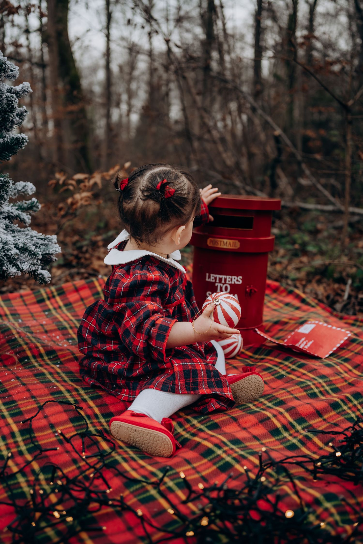 Child in plaid shirt lies on festive tartan blanket with red mailbox and Christmas lights in winter forest setting.
