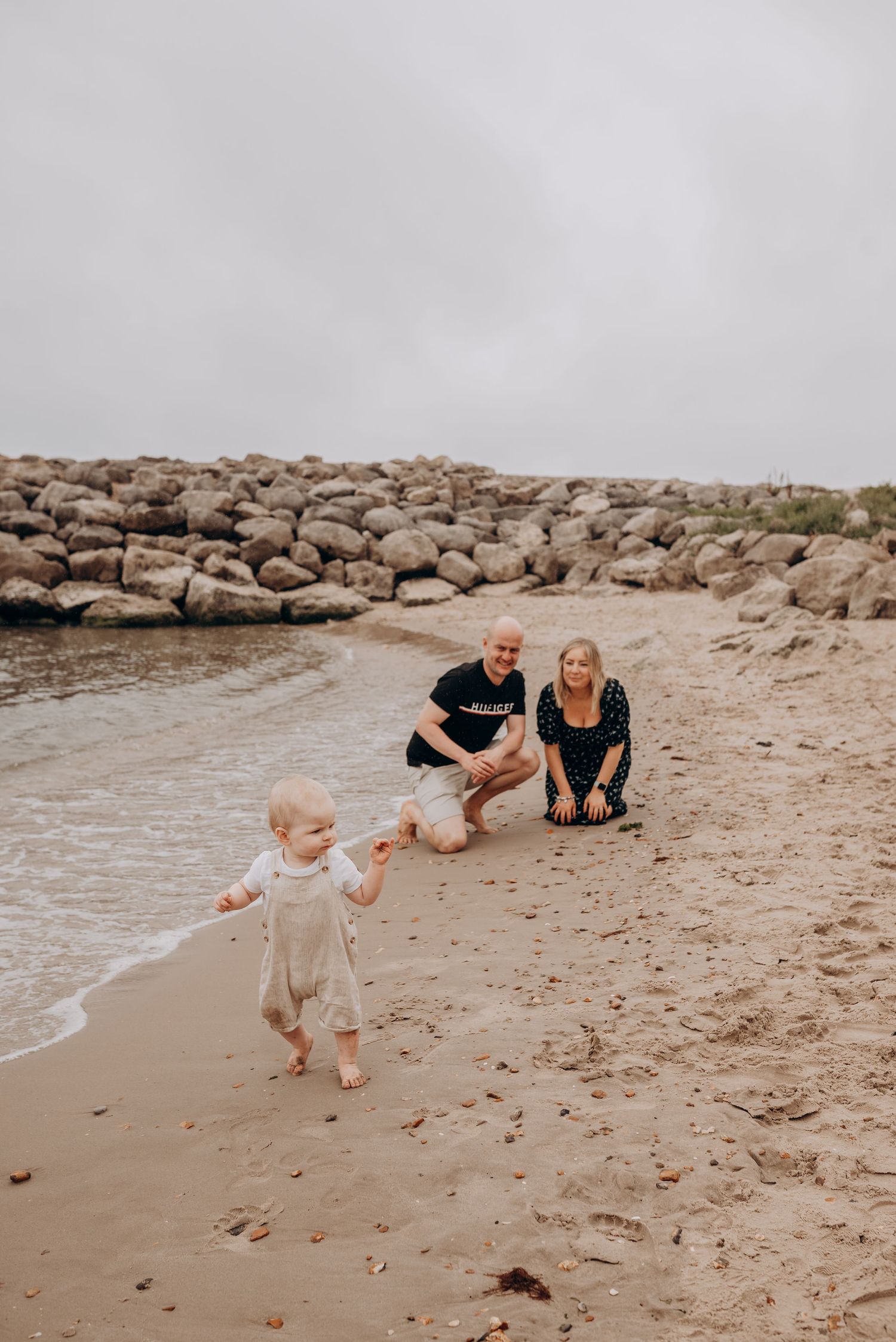Child plays near ocean shore while others watch from rocky beach.