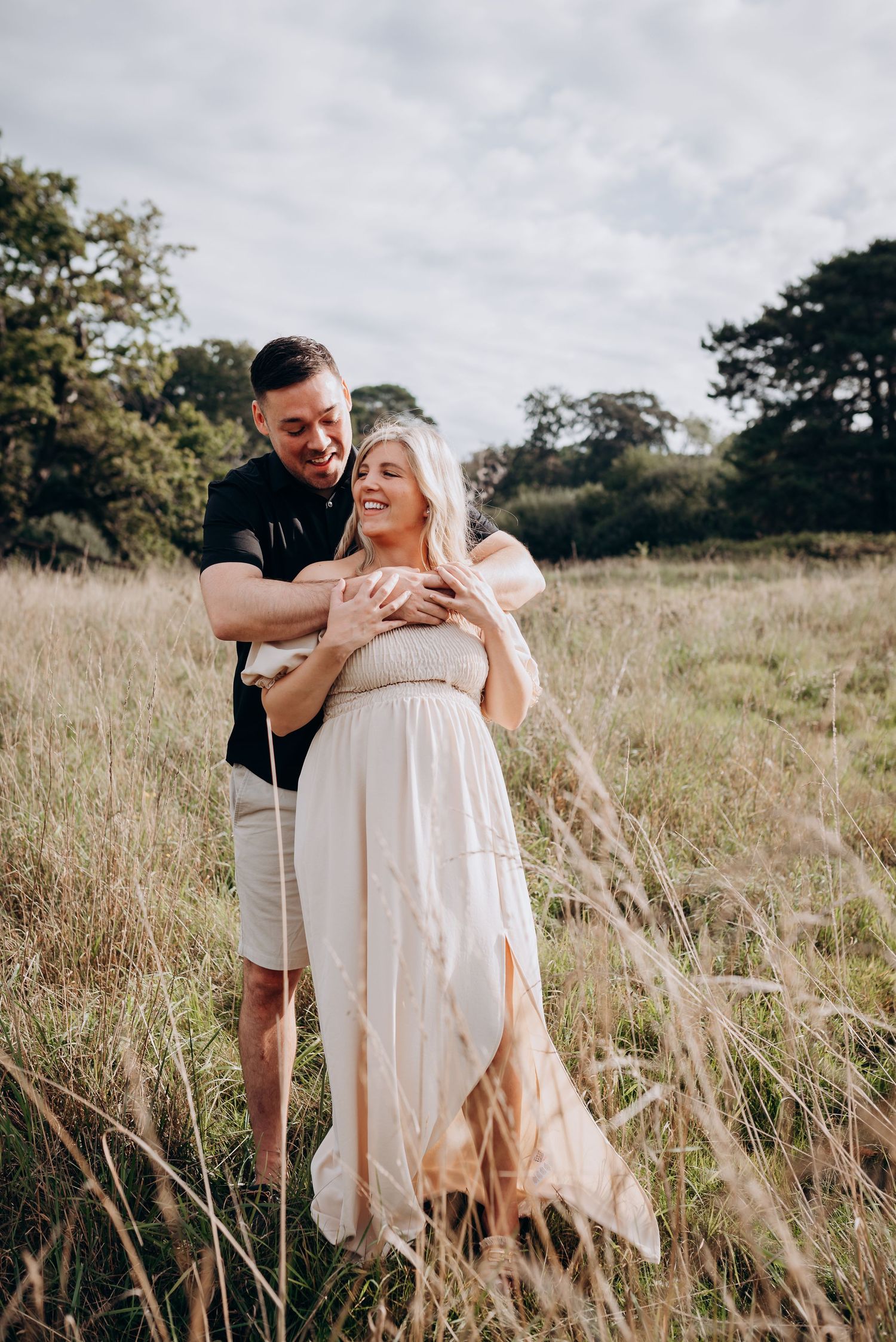 Romantic maternity photo of couple embracing in tall grass field during golden hour.