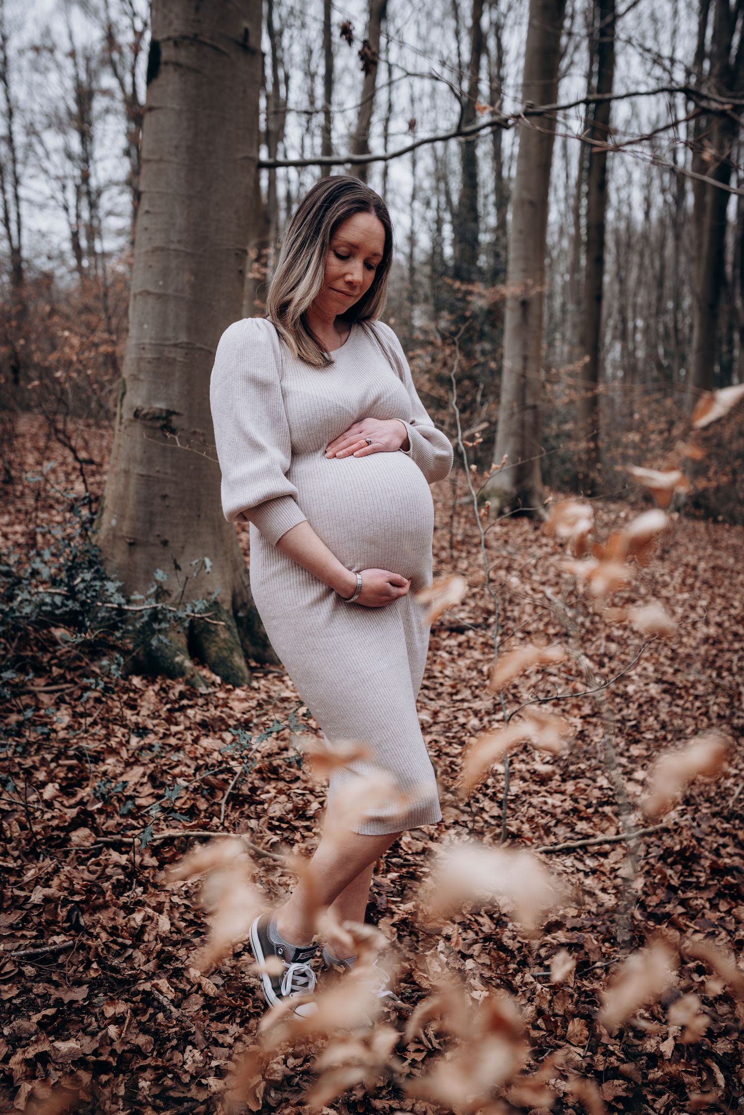 Pregnant woman in cream sweater dress poses thoughtfully in autumn forest setting.