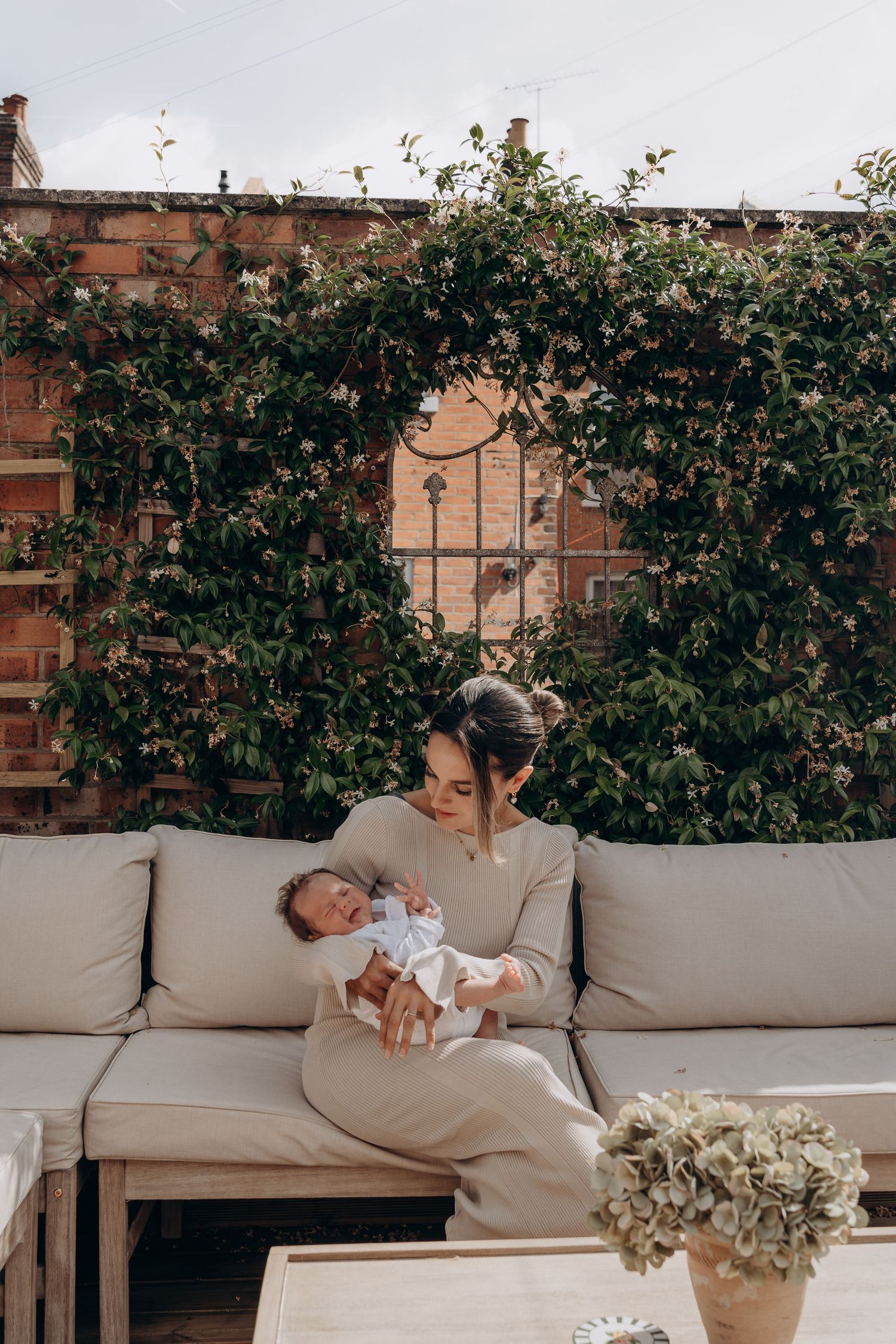 A mother sits on an outdoor sofa holding her newborn baby, surrounded by lush climbing plants on a brick wall.