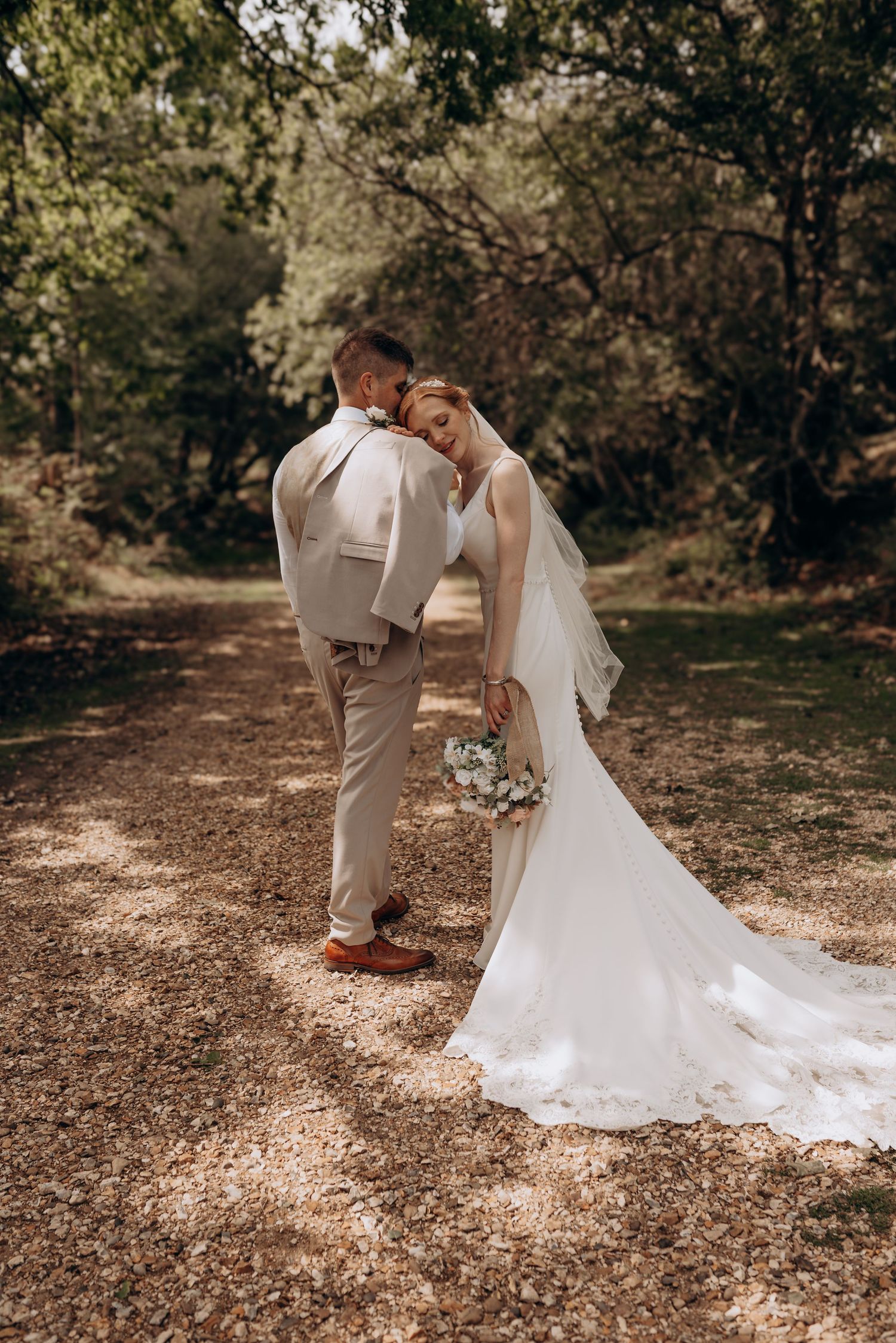 A romantic wedding photo of a couple sharing an intimate moment on a wooded dirt path, with the bride in a flowing white gown.