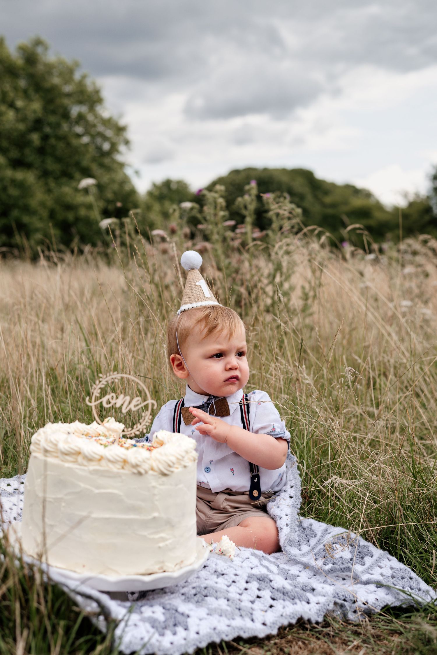 Baby in formal wear and party hat reaches for white birthday cake in tall grass setting.