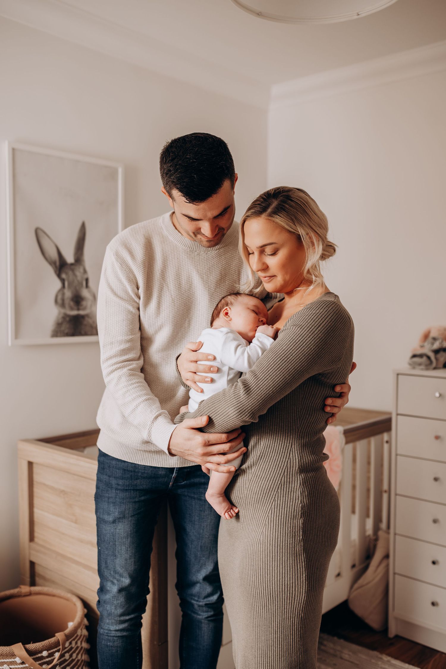 Parents in neutral tones hold and embrace their newborn baby in a minimalist nursery setting.