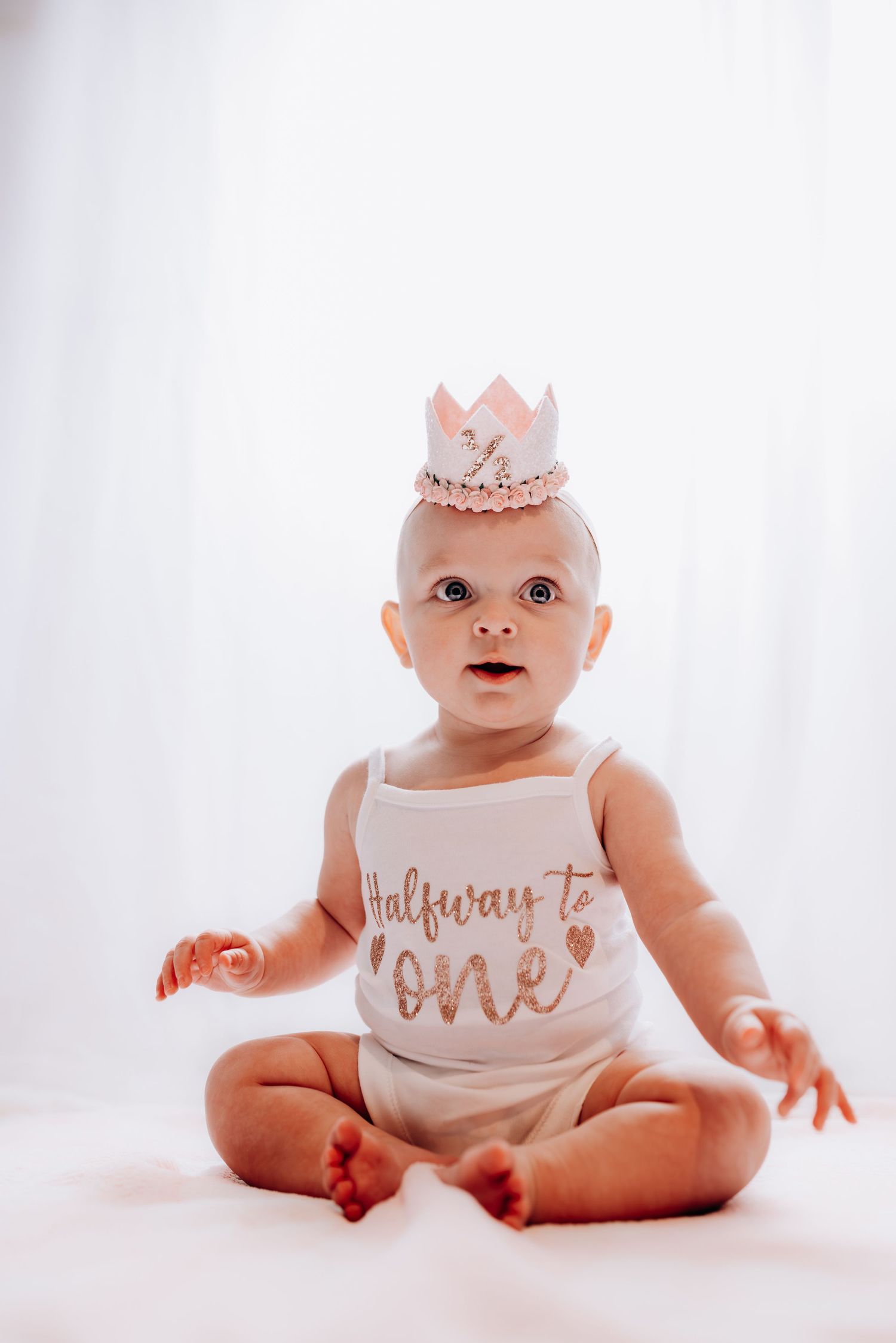 Baby in white outfit with crown sitting against white backdrop during milestone photoshoot.