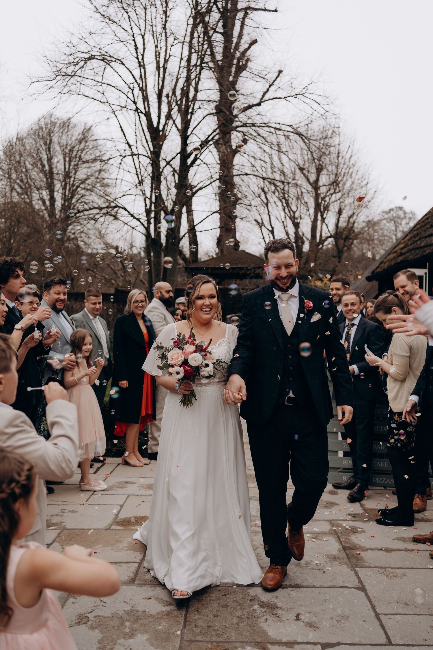 Newlyweds walk down stone path as wedding guests shower them with confetti on overcast winter day.
