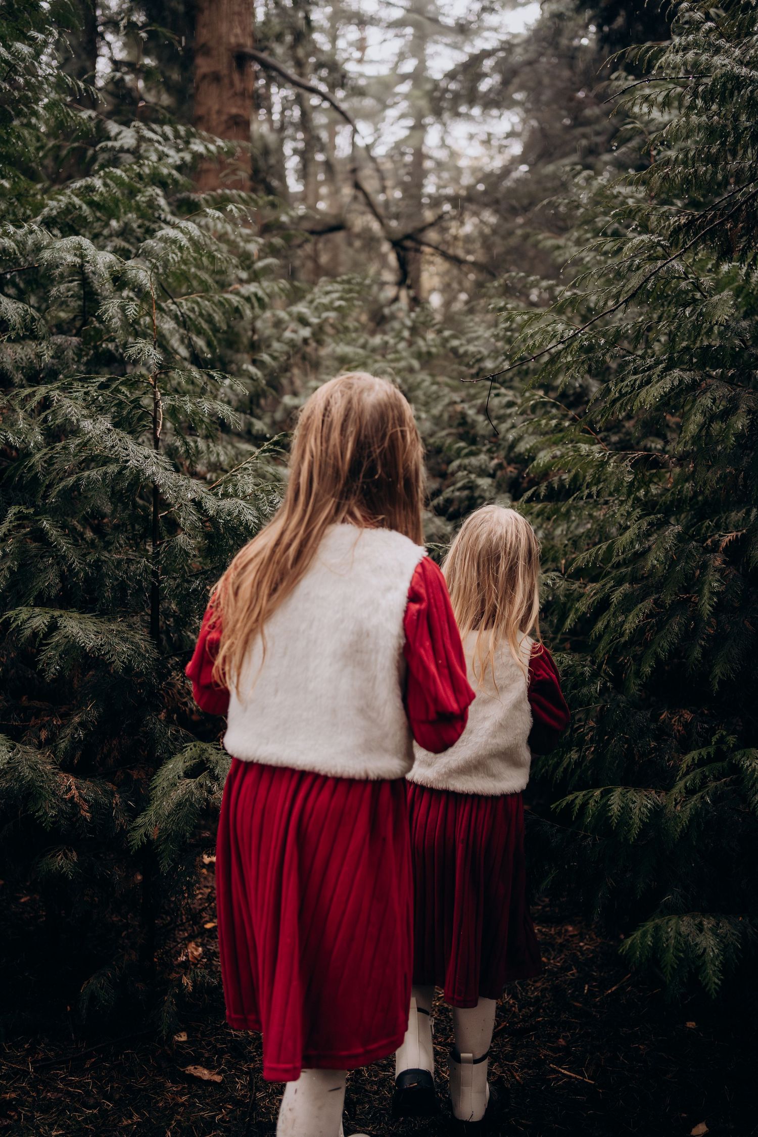 Two figures in matching red dresses and white fur vests walk through an evergreen forest path on a moody day.