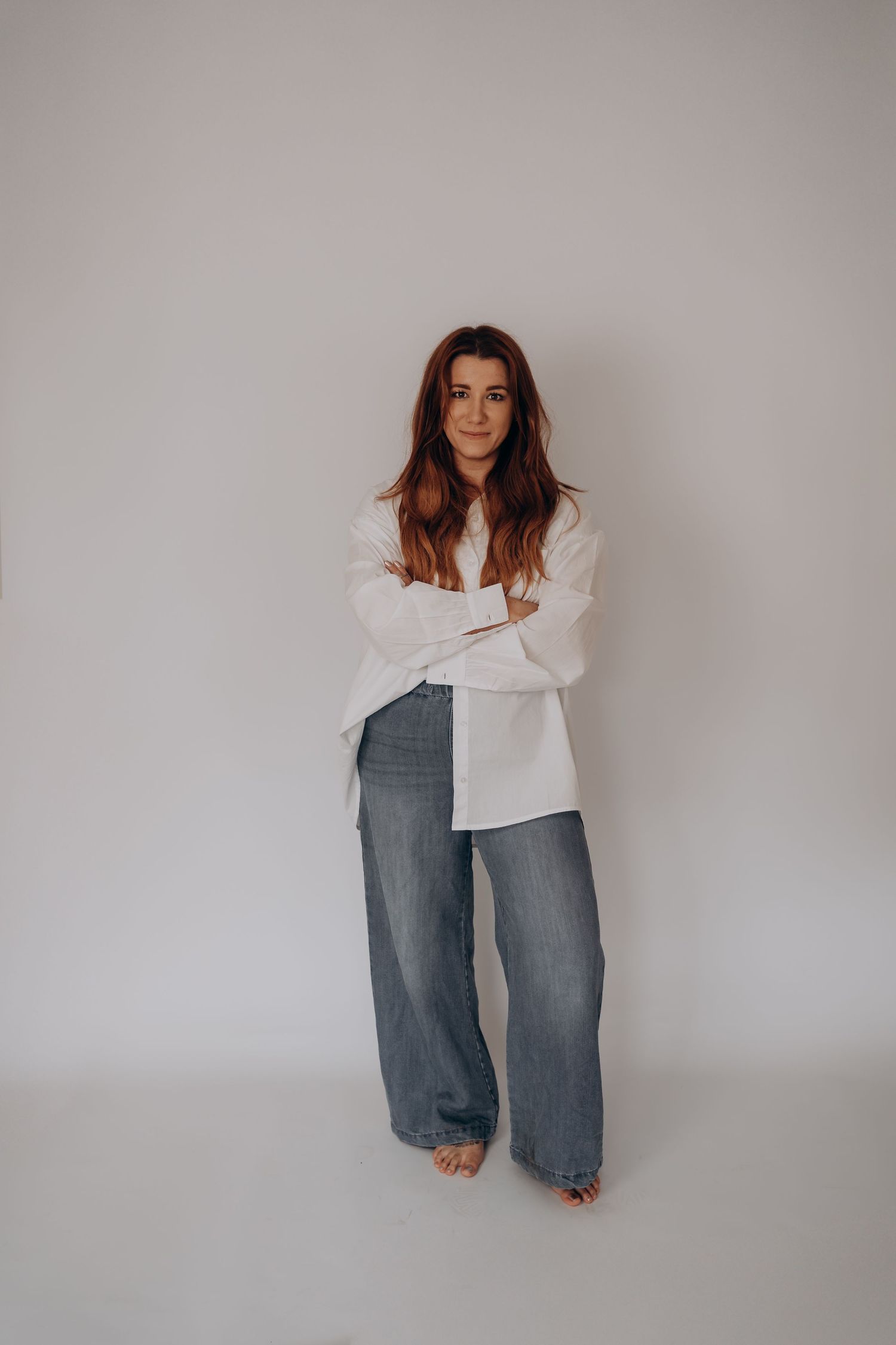 A model wearing a white sweater and wide-leg gray jeans poses against a neutral wall in a minimalist fashion shoot.