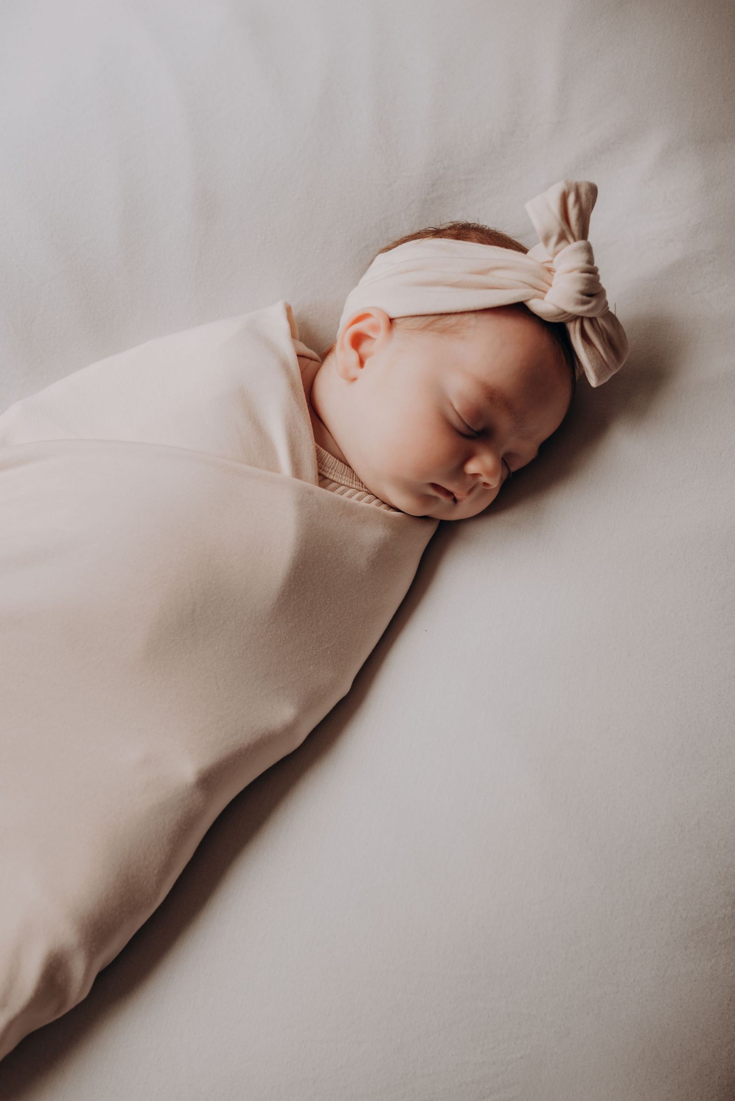 Sleeping baby in cream outfit rests peacefully on white bedding.