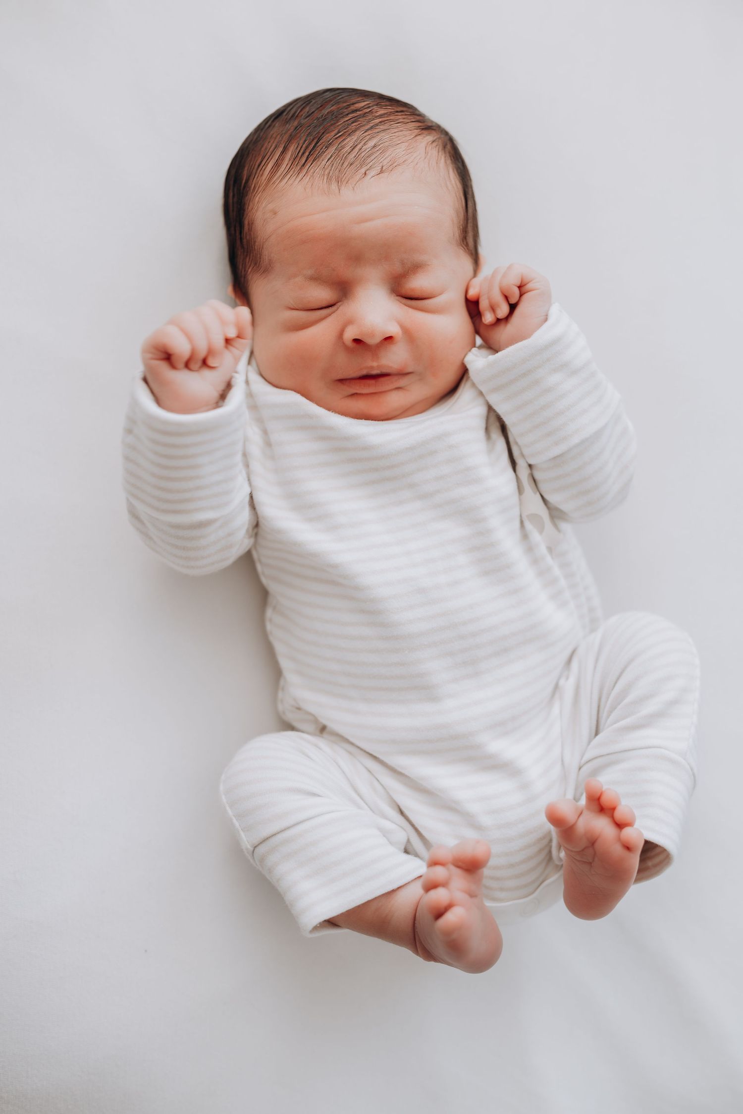 Newborn baby in striped white onesie stretching and yawning on white background.