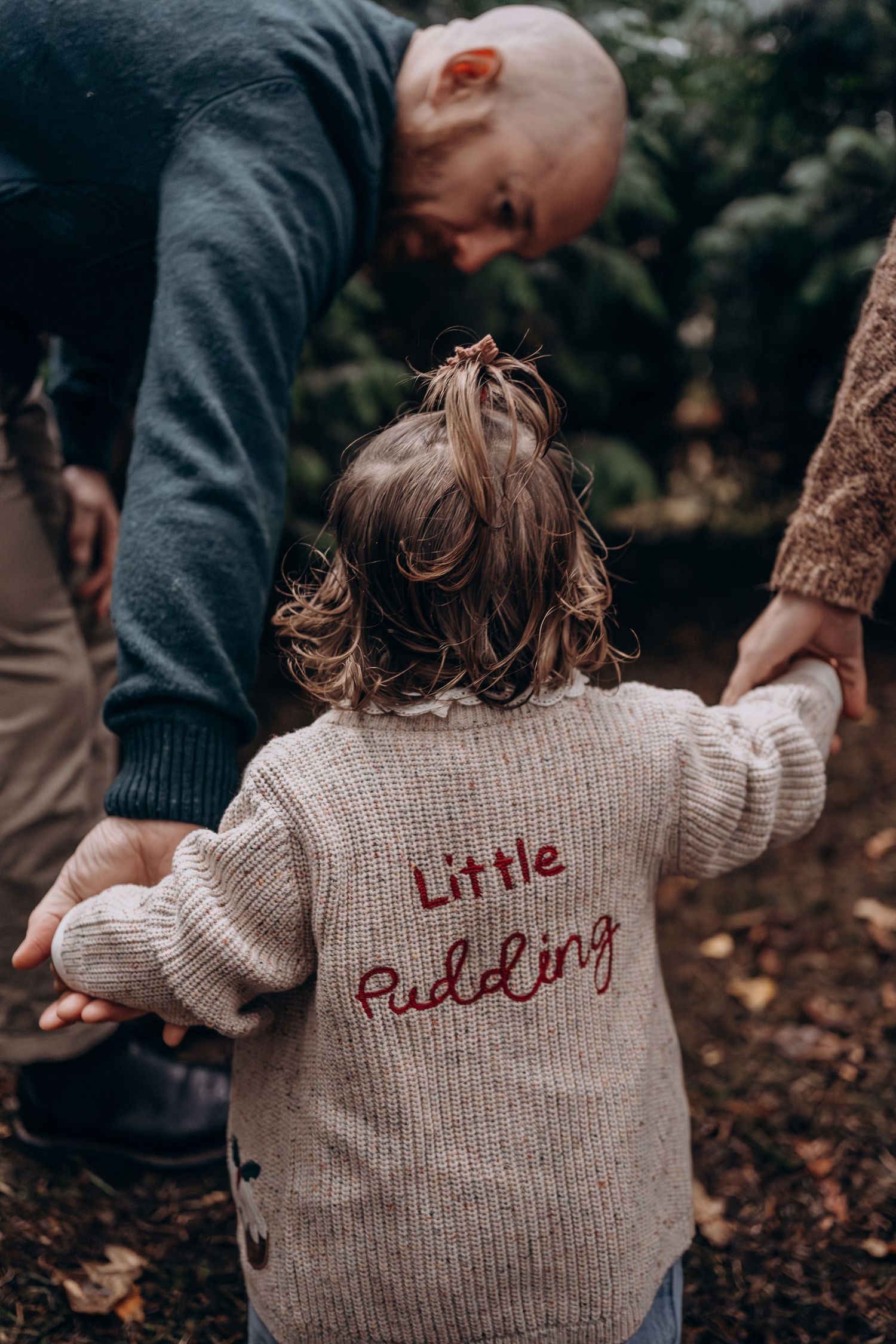 A child in a beige knit sweater with text embroidered on the back plays outdoors in an autumn setting.