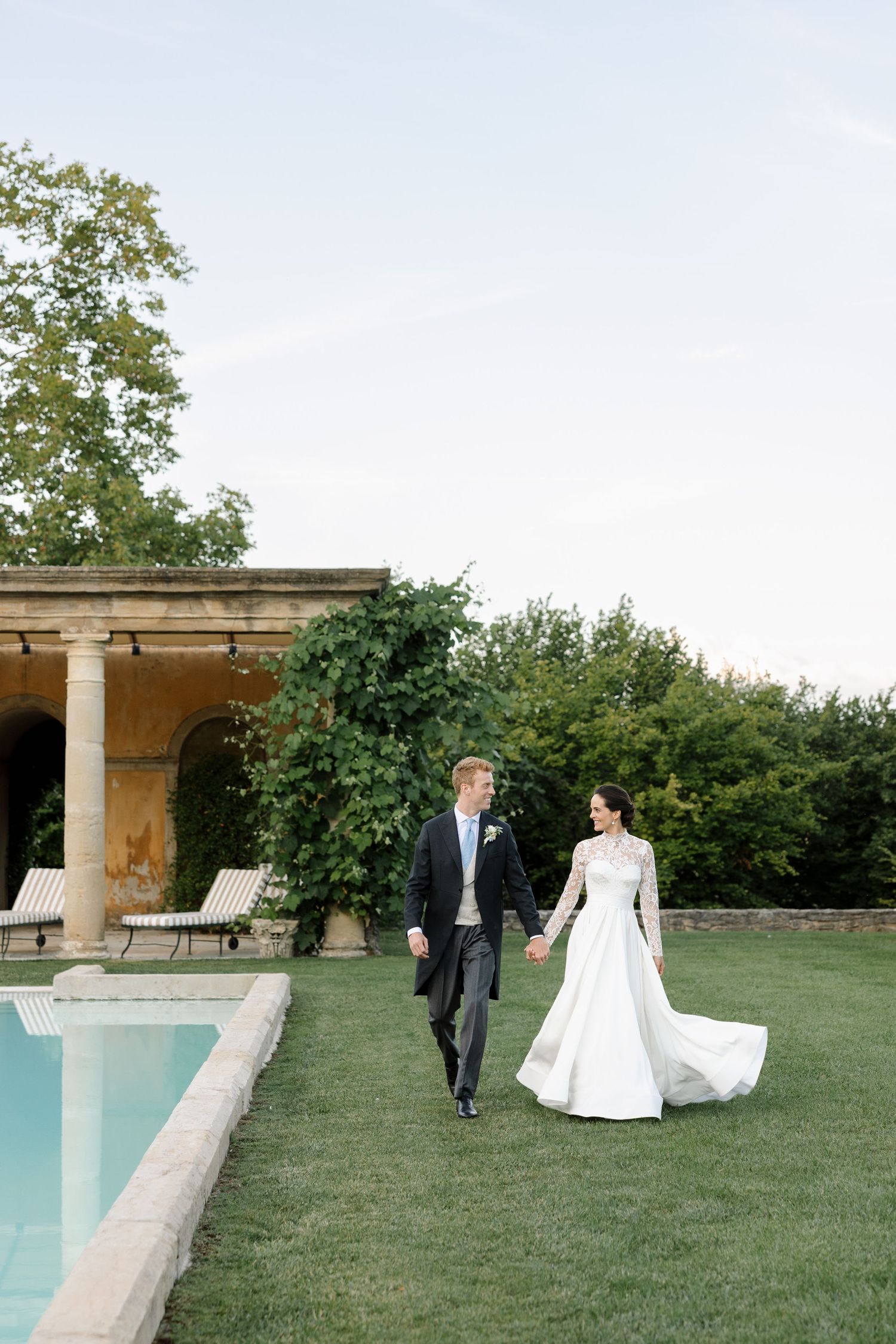 A wedding couple walks near a classical pool terrace surrounded by manicured gardens.