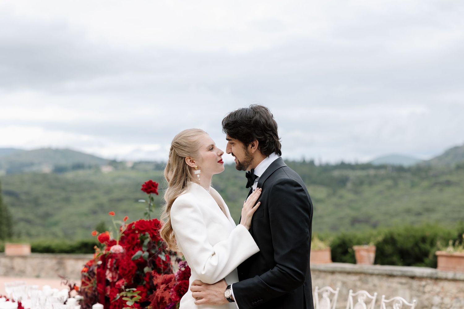 A romantic wedding couple embraces against a scenic mountain backdrop with red roses in the foreground.