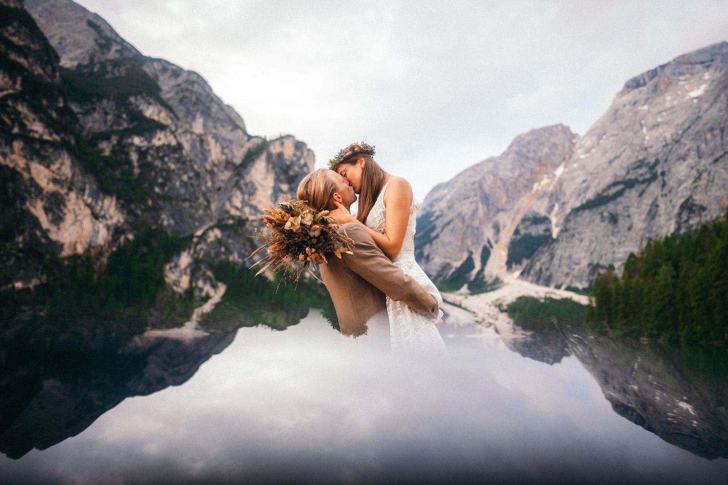 Wedding couple embracing and kissing by a serene mountain lake with stunning reflections in the Dolomites.