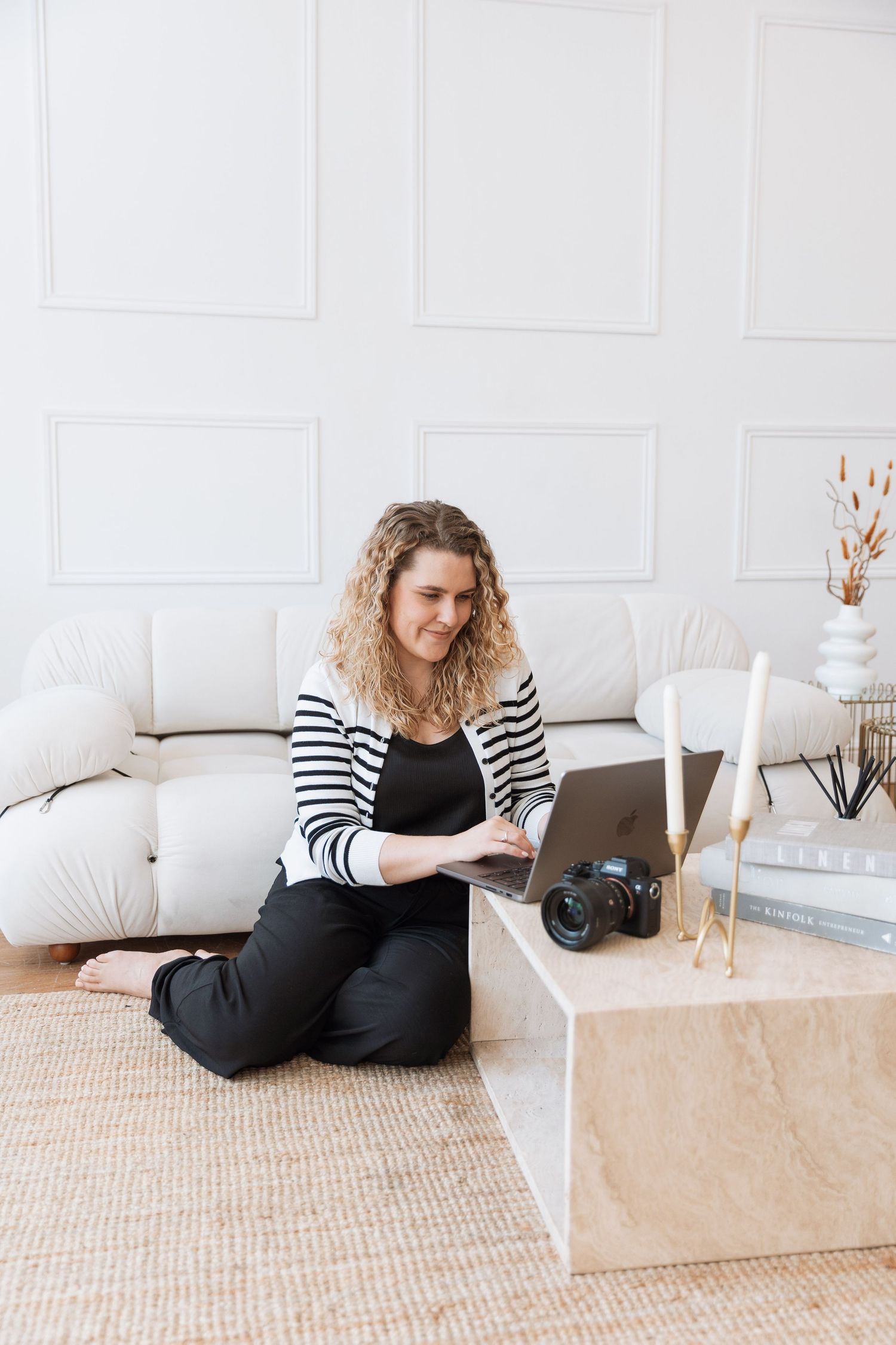 Someone in a striped cardigan works on a laptop while sitting on the floor with camera equipment and decor nearby.