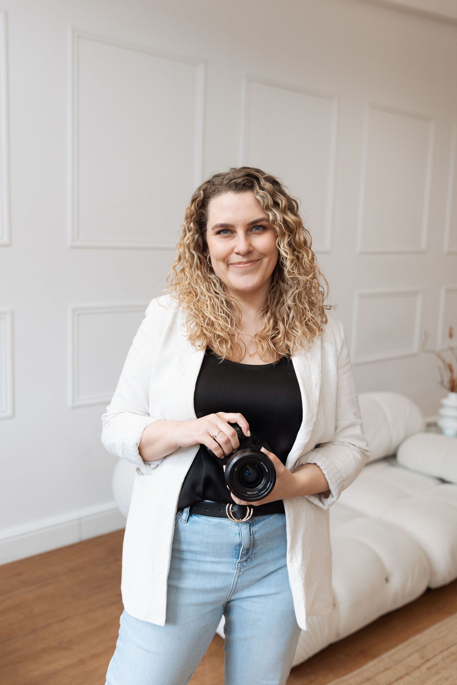 Professional photographer stands smiling with camera in a bright modern bedroom with white walls.