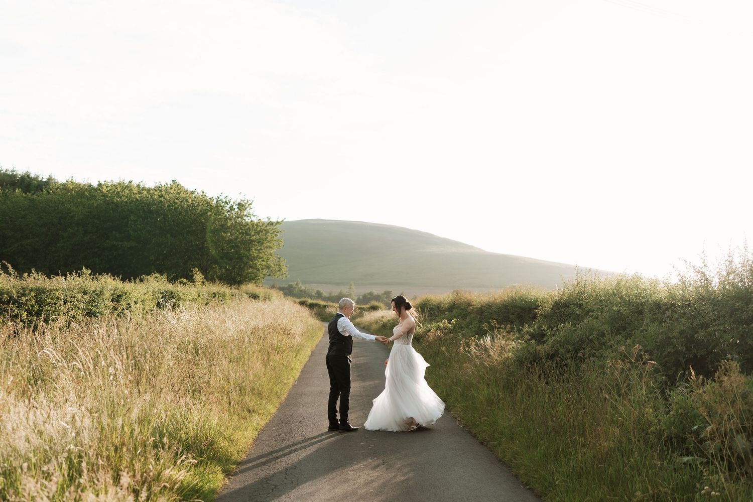 A scenic country road winds through rolling hills with tall grass and wildflowers at sunset.