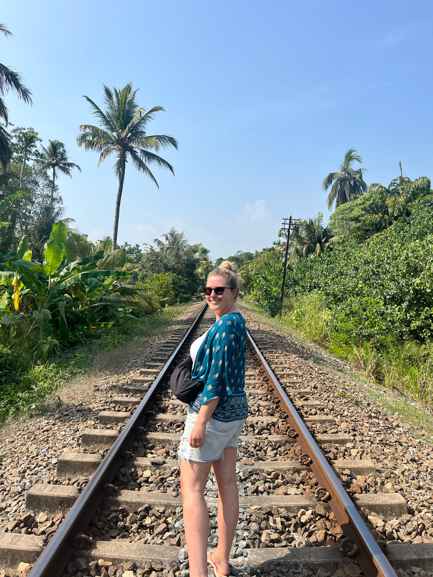 Person standing on tropical railway tracks surrounded by palm trees and lush vegetation in a scenic setting.