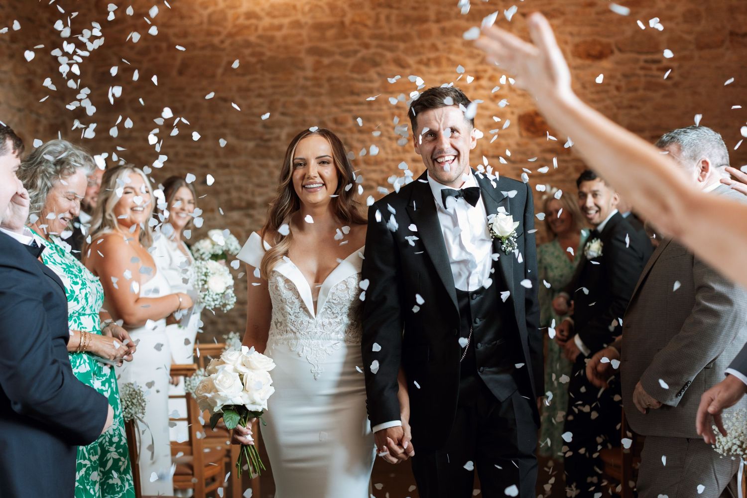 Newlyweds walk through shower of white confetti during wedding celebration with guests on wooden background.
