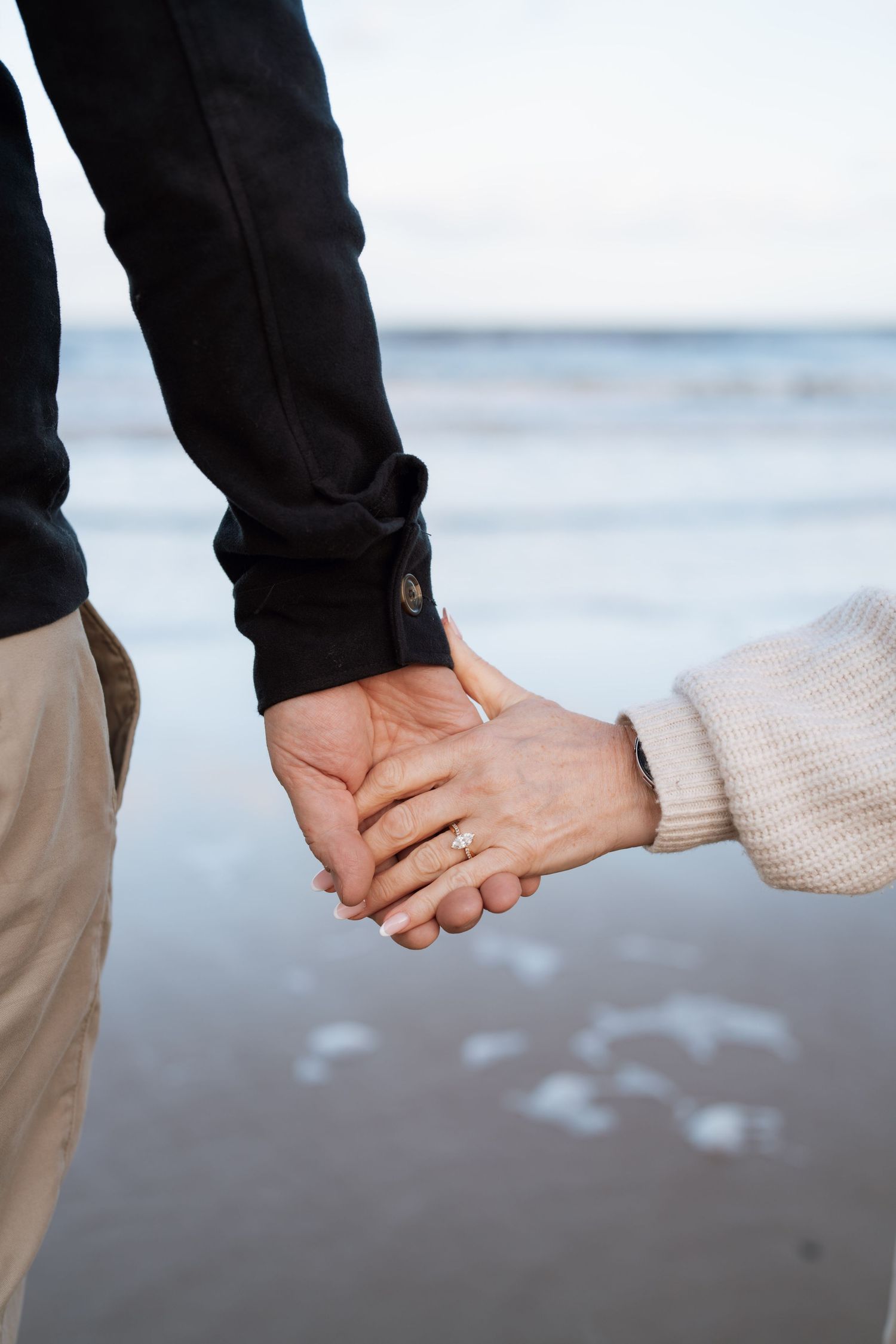 Close-up of two people holding hands on a beach, one wearing black pants and one in a cream sweater.