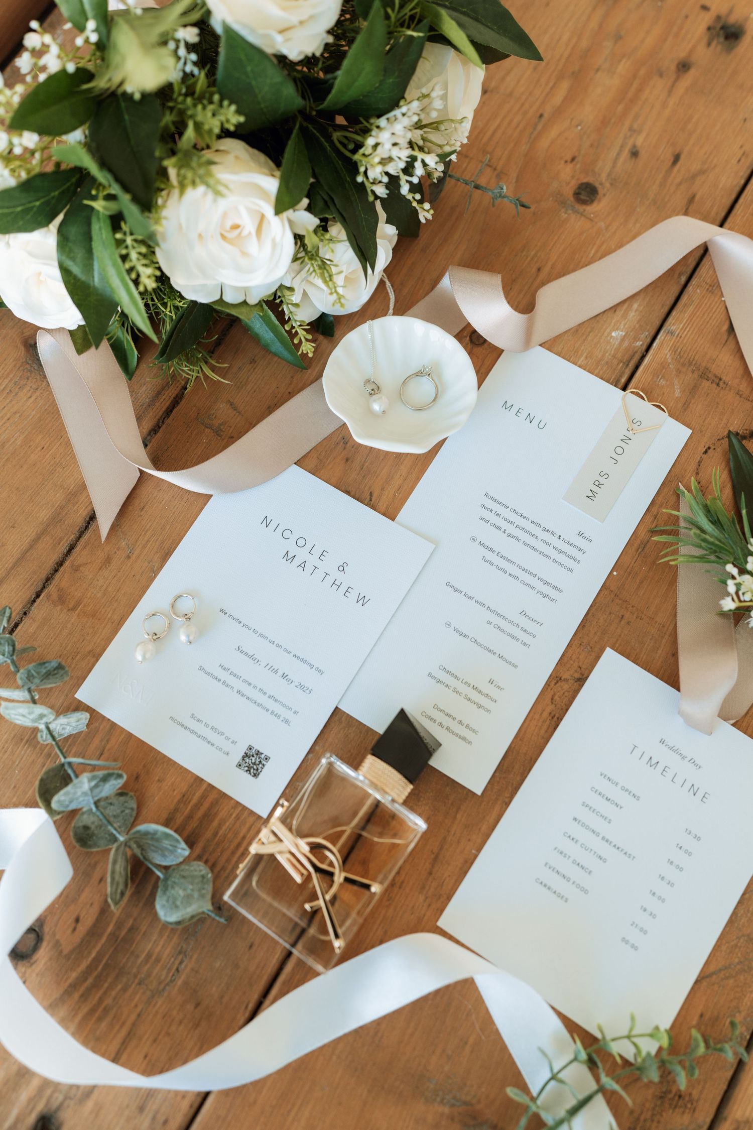 Wedding stationery and floral arrangements displayed on rustic wooden table with white roses and eucalyptus leaves.