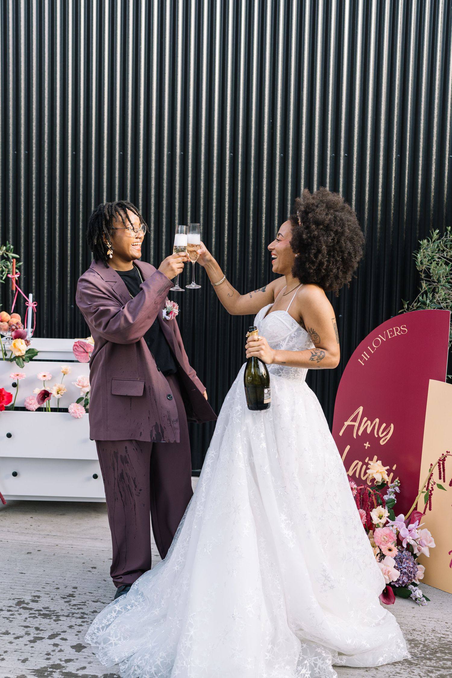 Wedding party in burgundy suits and white dresses toasting with champagne glasses against a black wall.