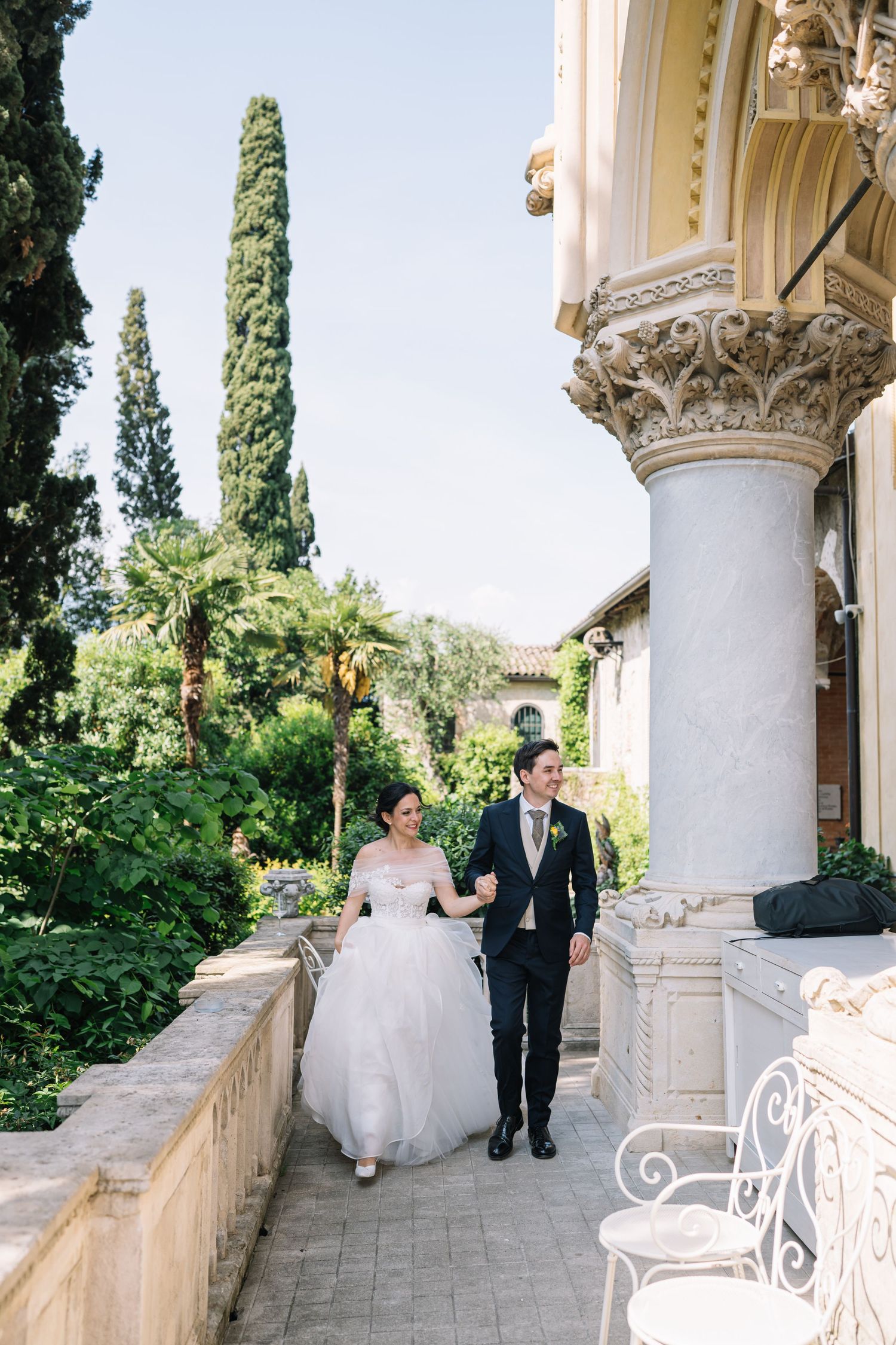 Elegant wedding couple strolls along a Mediterranean villa's columned terrace with cypress trees in the background.