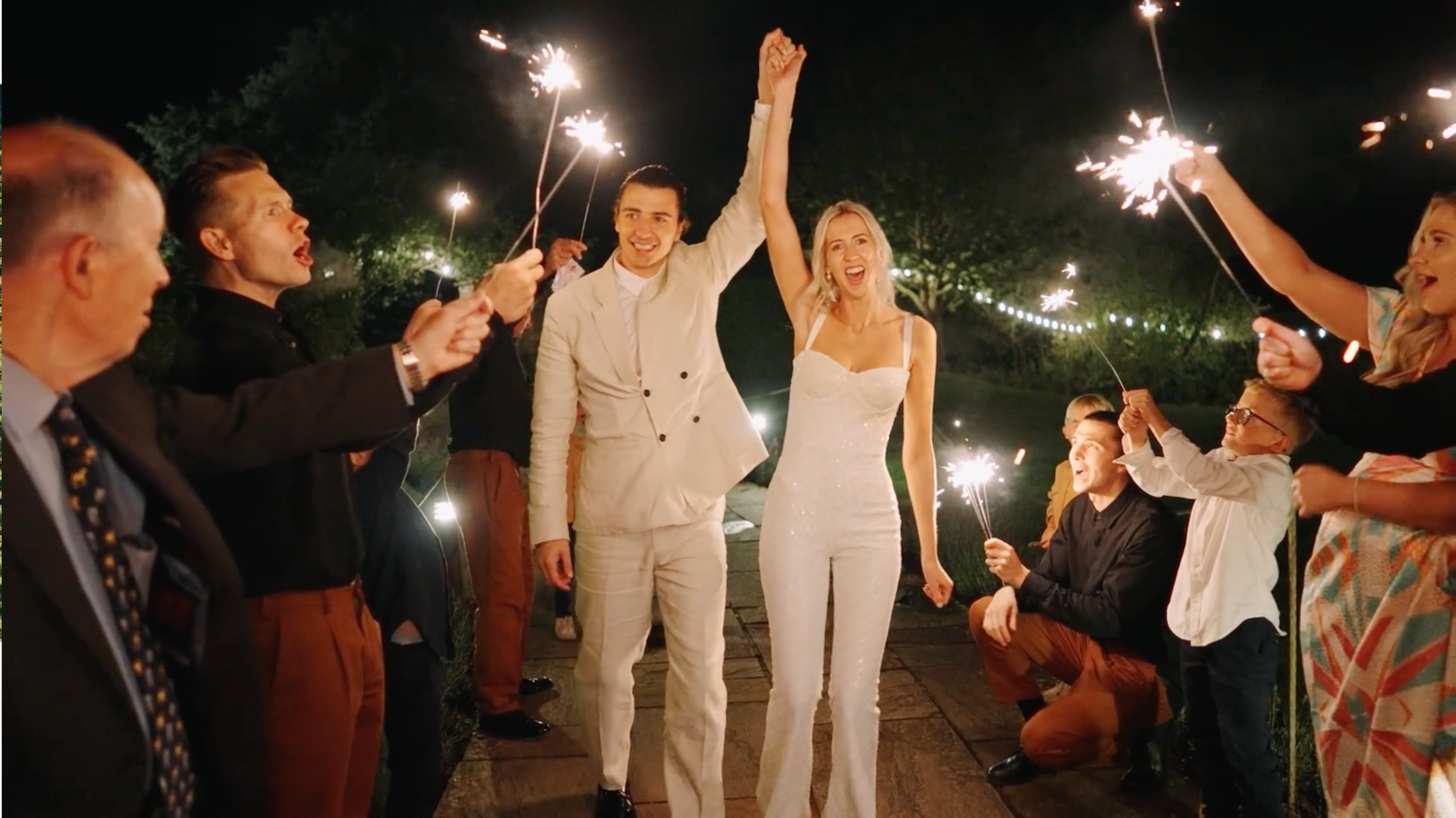 Newlyweds in matching white outfits walk through a sparkler exit line during nighttime wedding celebration.