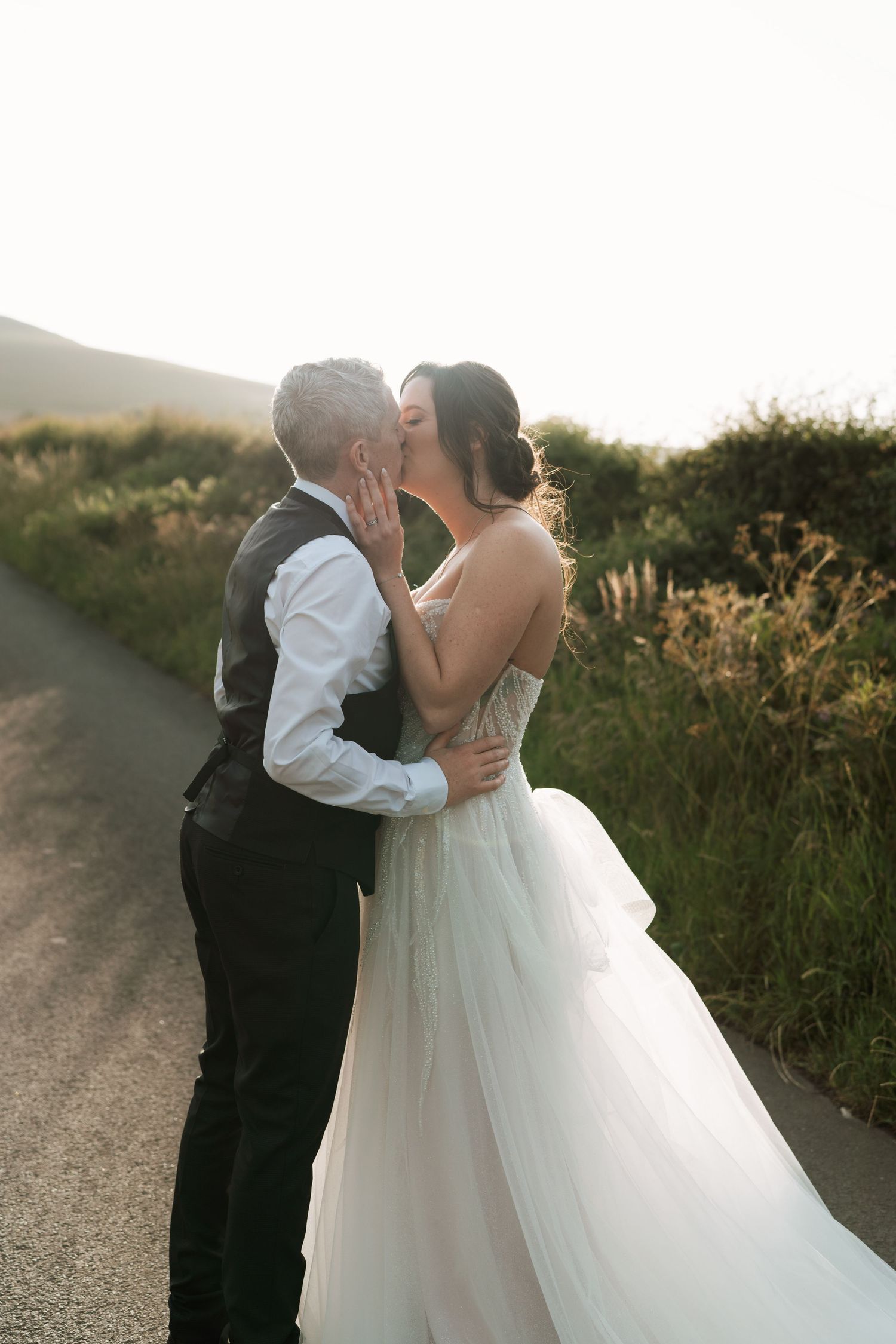 A romantic wedding couple embraces on a scenic path at sunset surrounded by golden wildflowers.