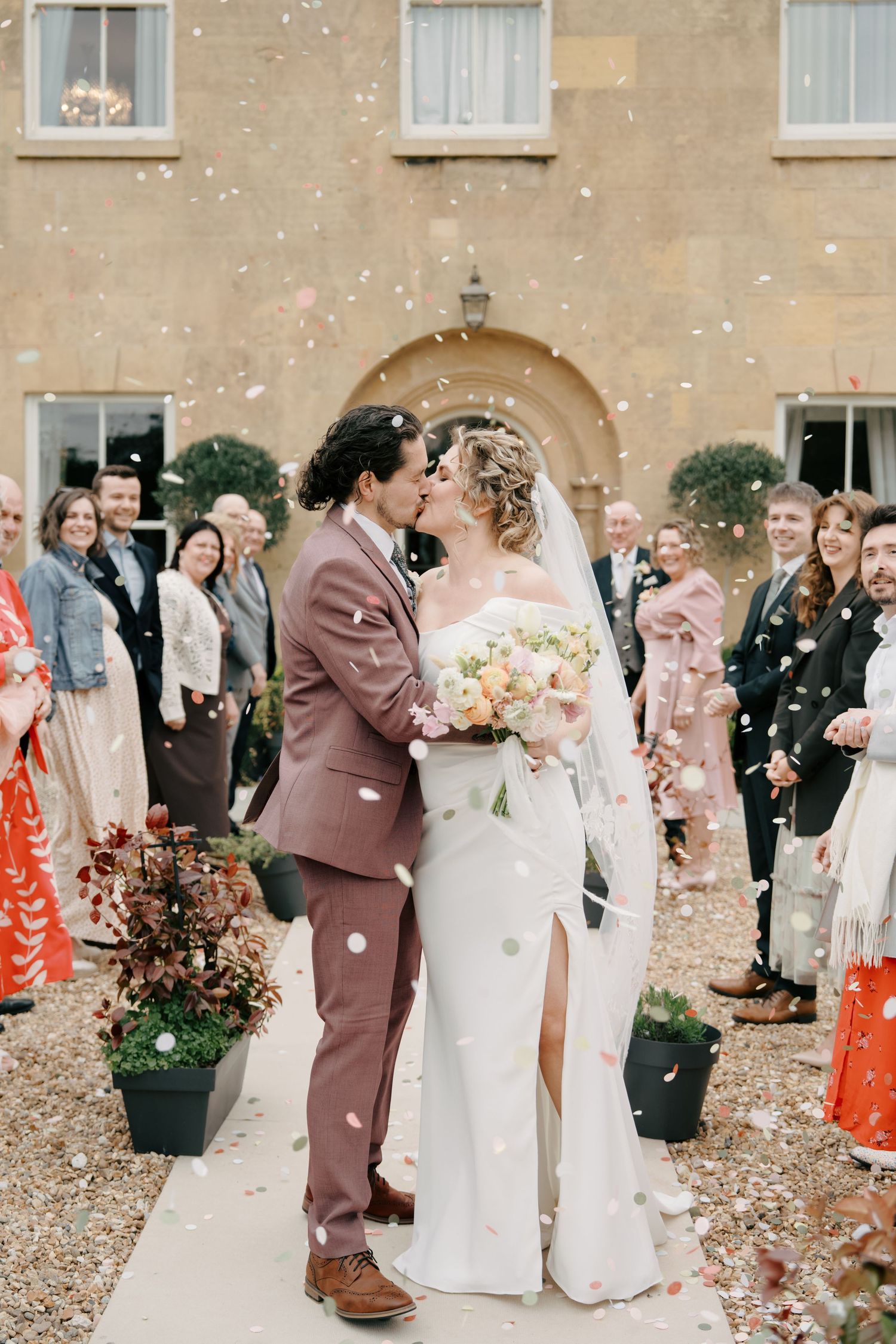 Newlyweds share a kiss as confetti falls around them outside a stone venue, surrounded by smiling wedding guests.