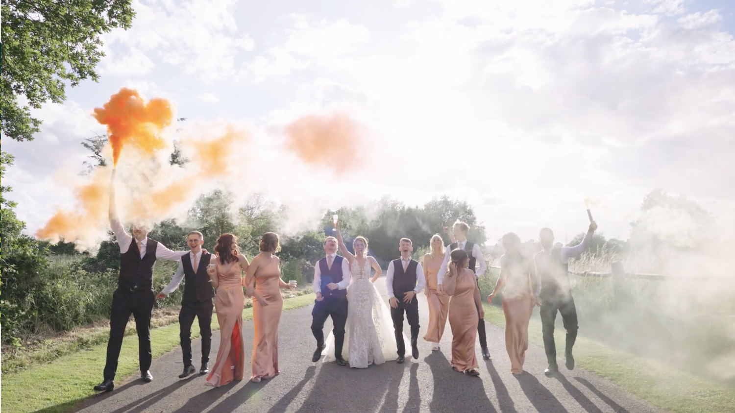 A wedding party poses in a row outdoors while orange smoke bombs create a dramatic atmospheric effect in the background.