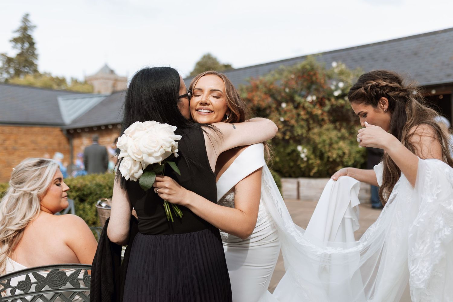 A joyful group wearing white dresses embrace and laugh together during an outdoor celebration.