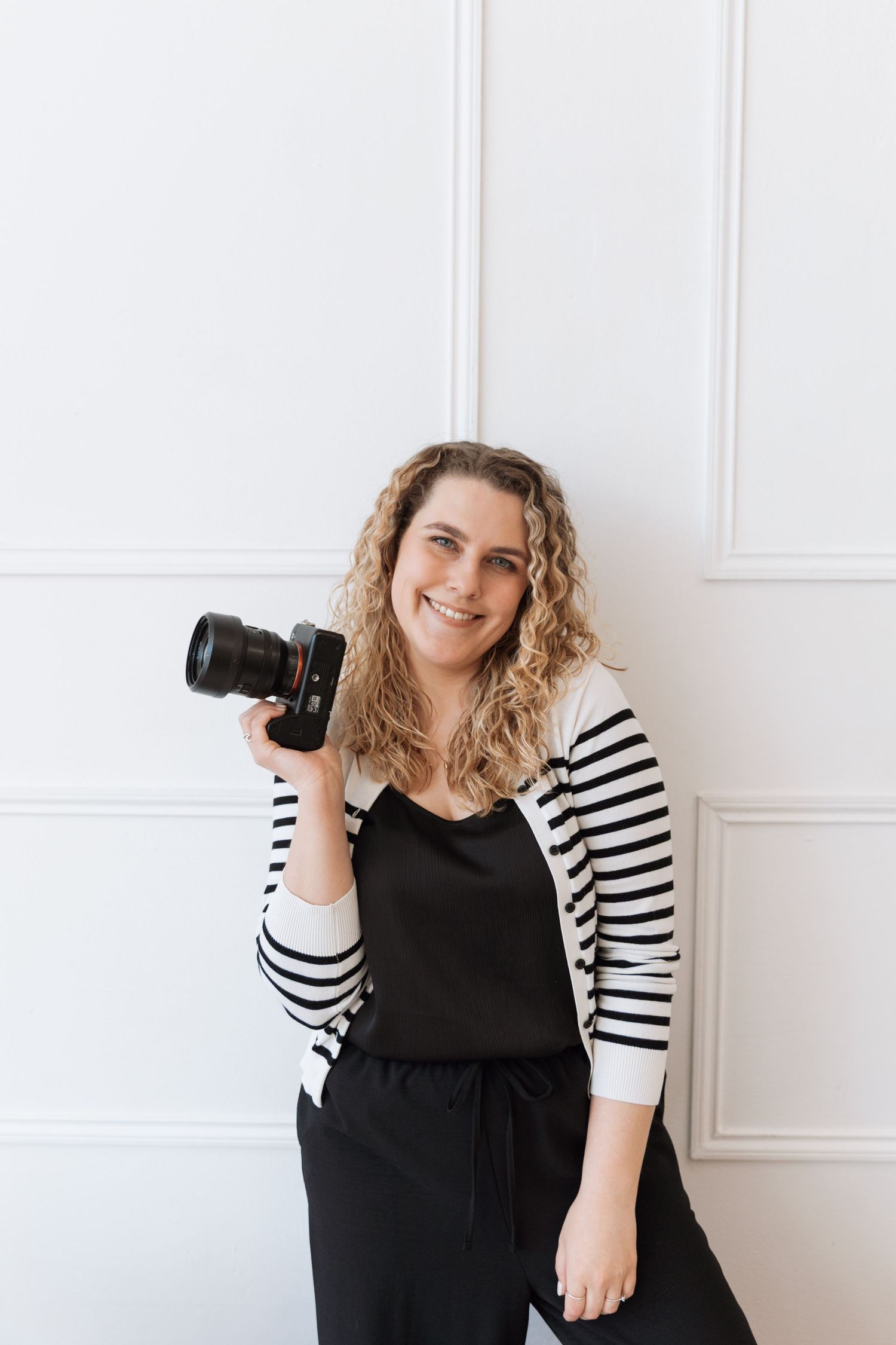 Professional photographer in black dress and striped cardigan poses with camera against white wall.