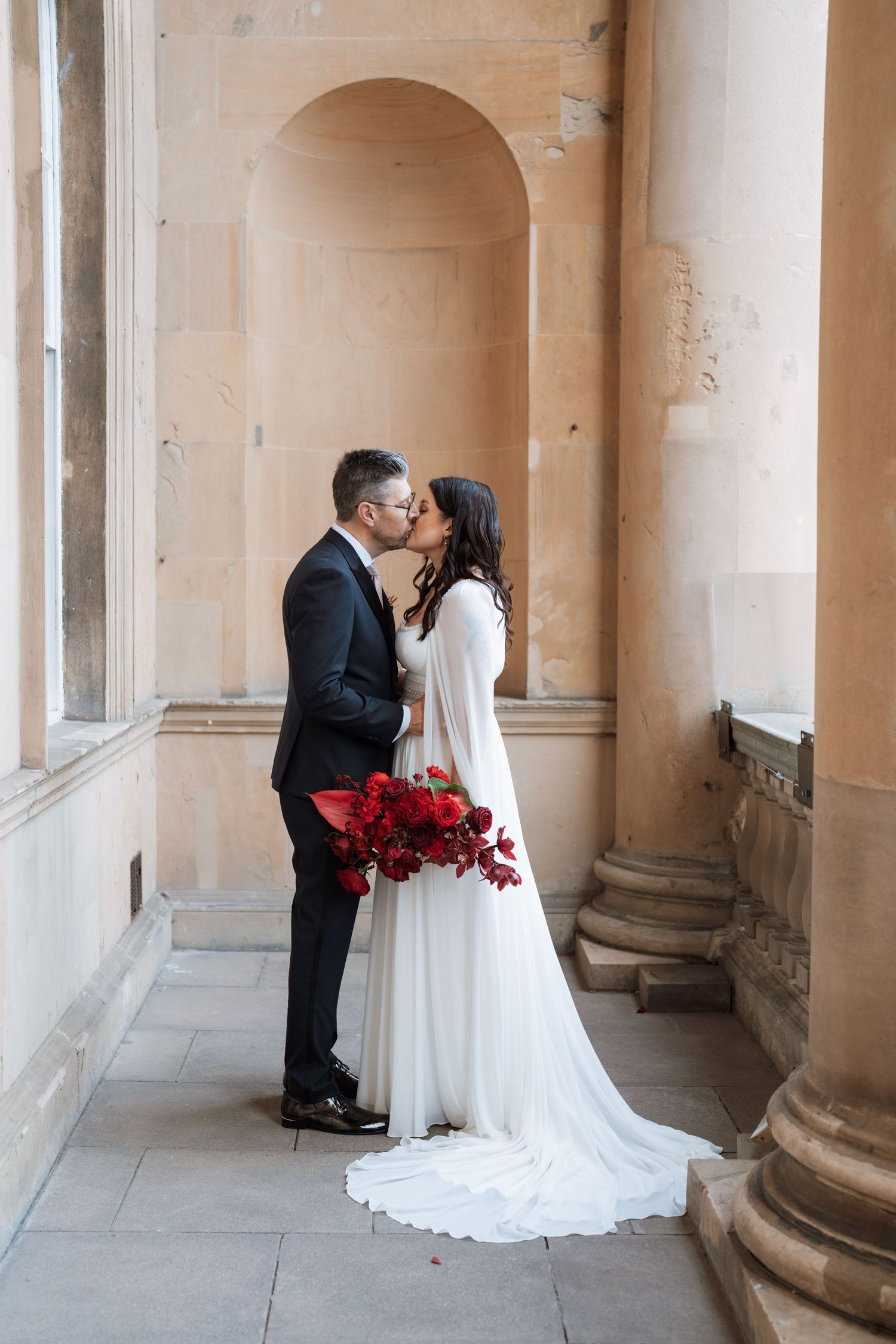Romantic wedding couple sharing intimate moments in an elegant colonnade with red floral accents.