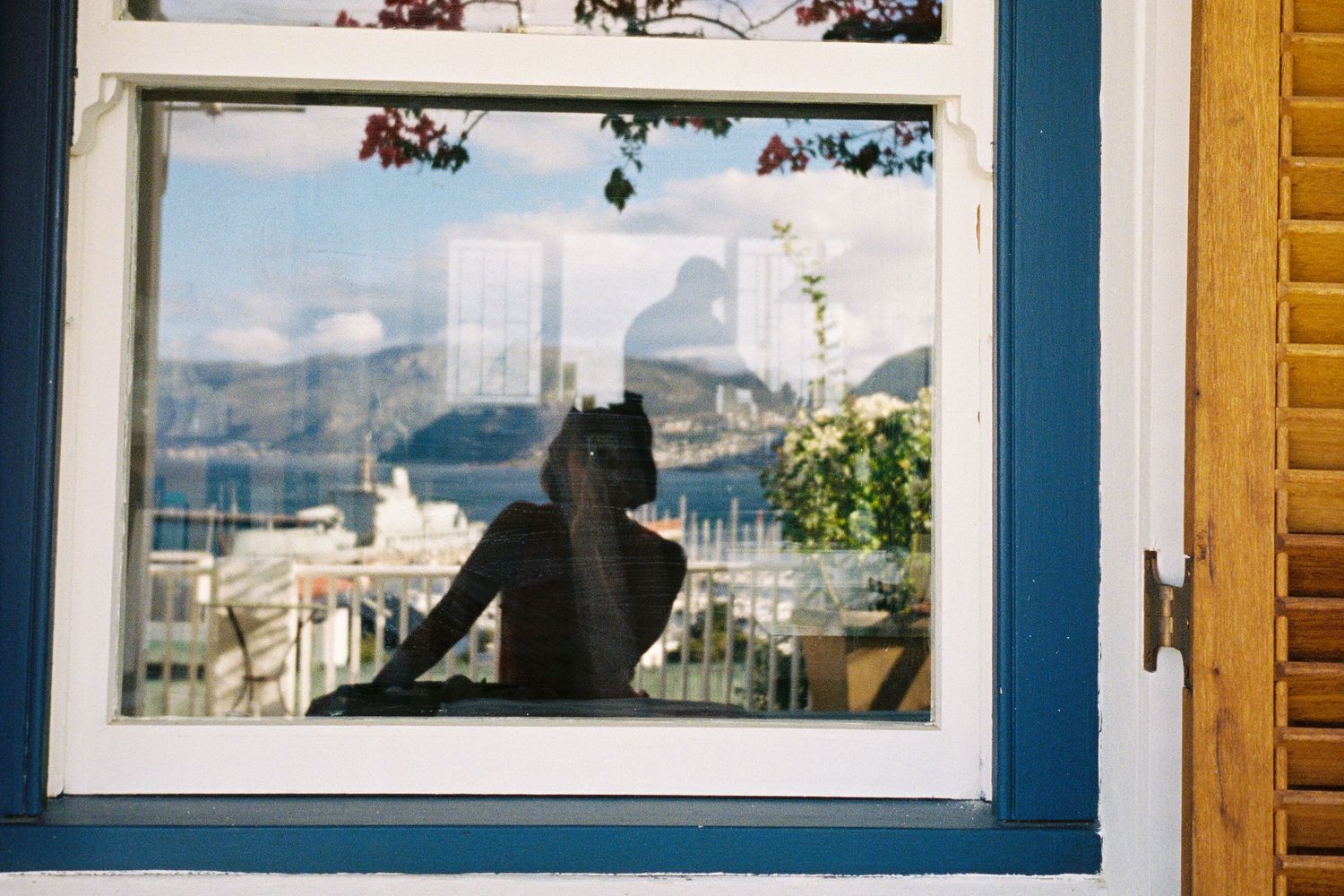 Self portrait of the artist in a window reflecting the ocean and mountains and boats in the back ground and also a person through the window