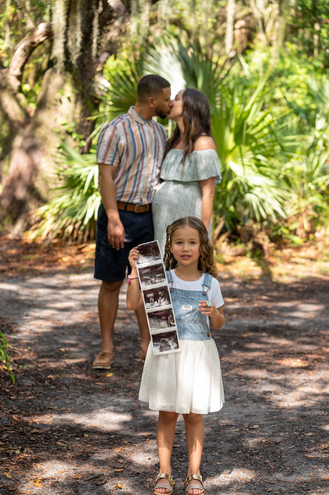 Child holds ultrasound photos while parents share tender moment in wooded background.