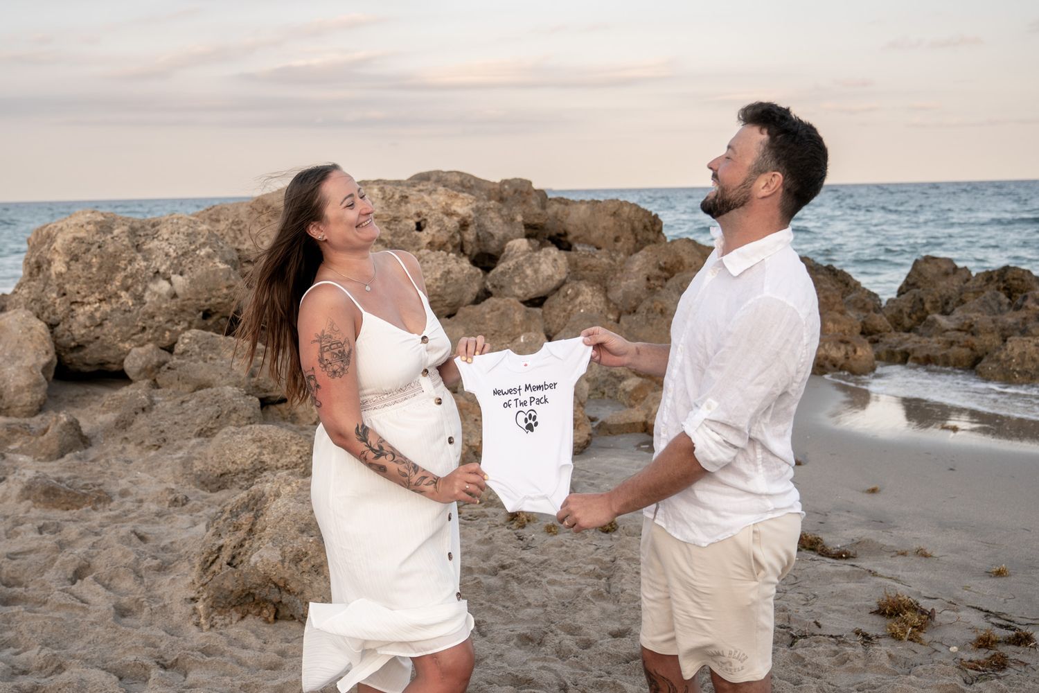 Couple on rocky beach holding baby onesie during pregnancy announcement photoshoot.