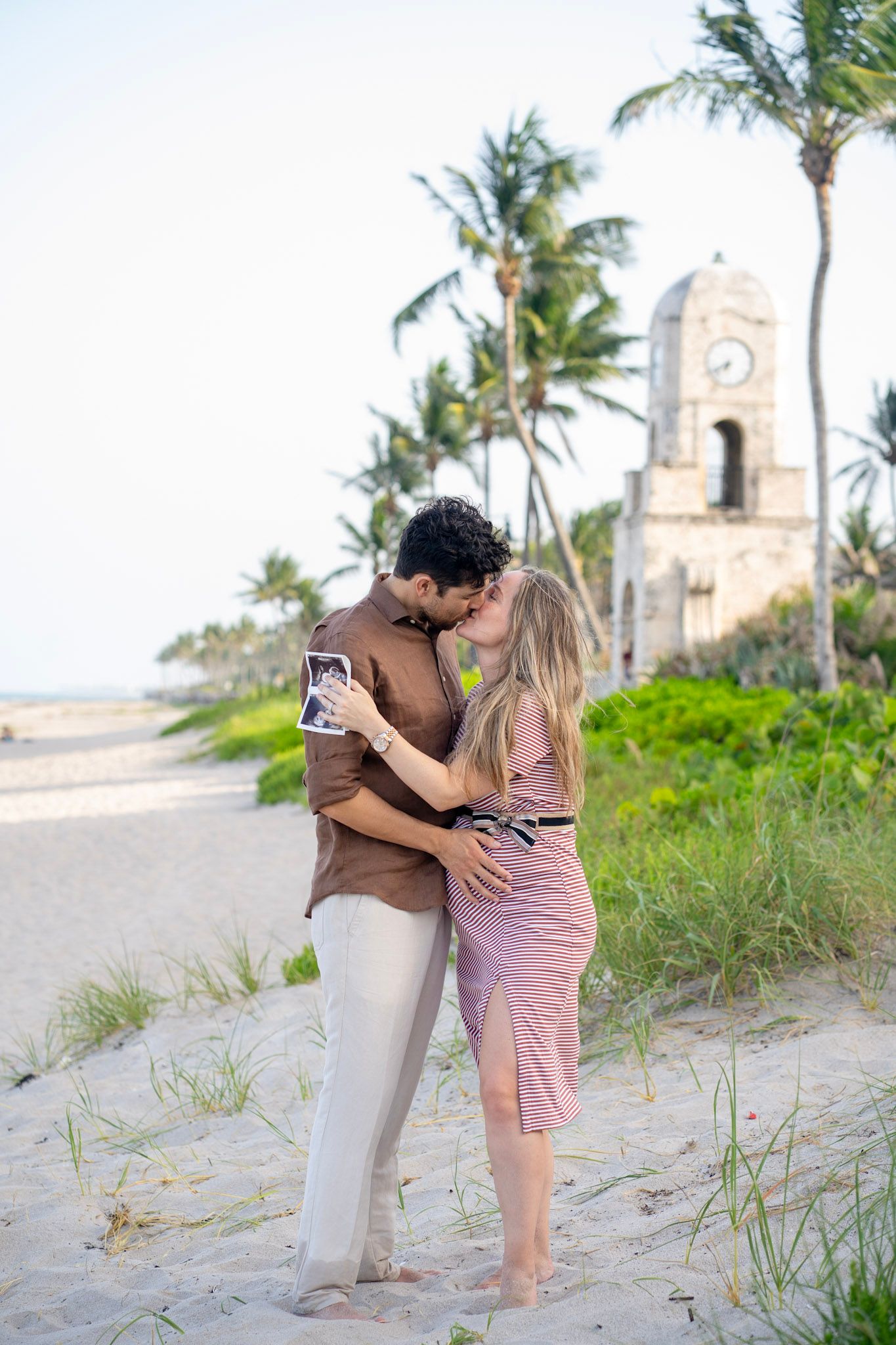 Romantic couple embracing on tropical beach with historic clock tower and palm trees.